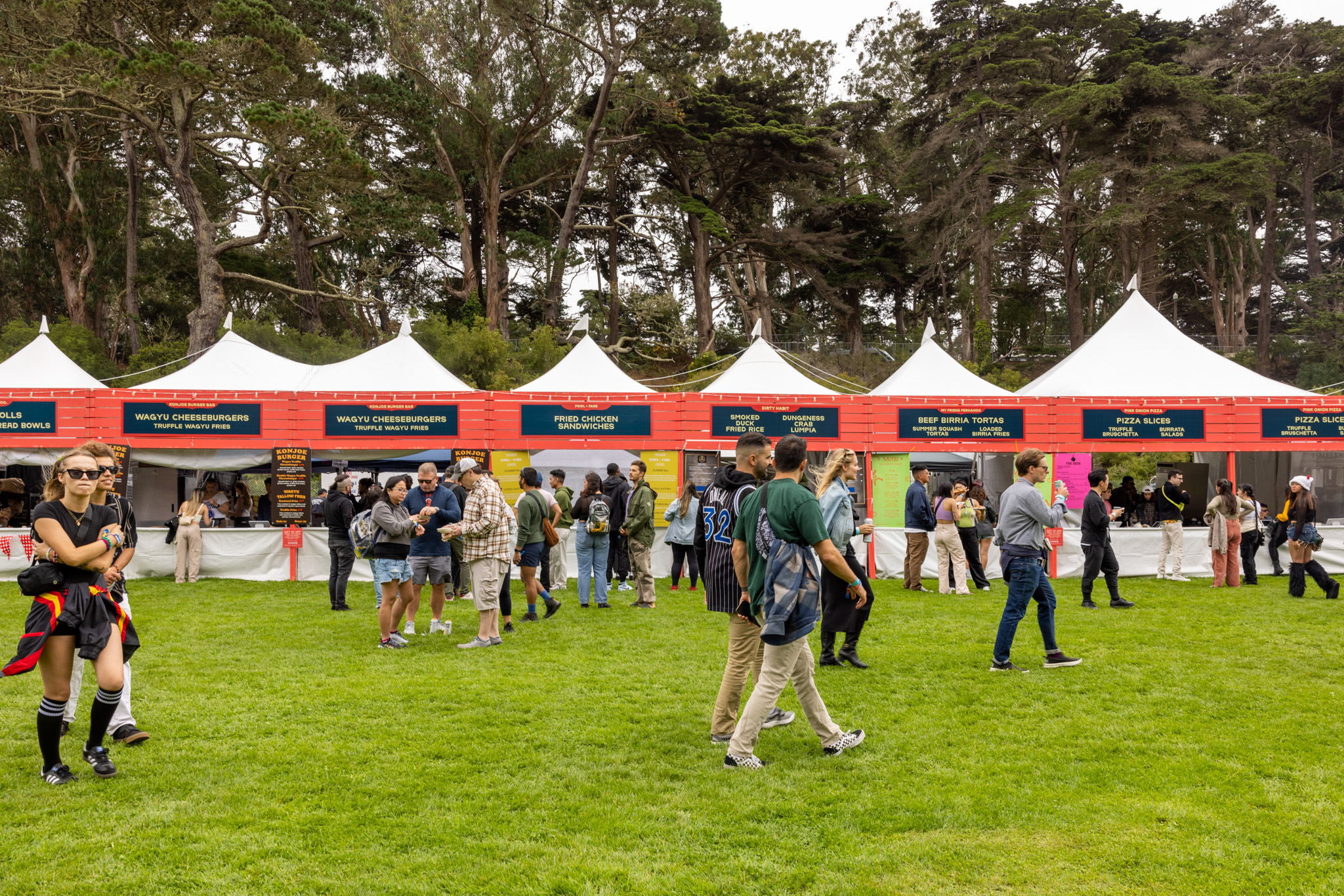 People are walking and queuing in front of various food stalls with signs under white tents at an outdoor event surrounded by tall trees.