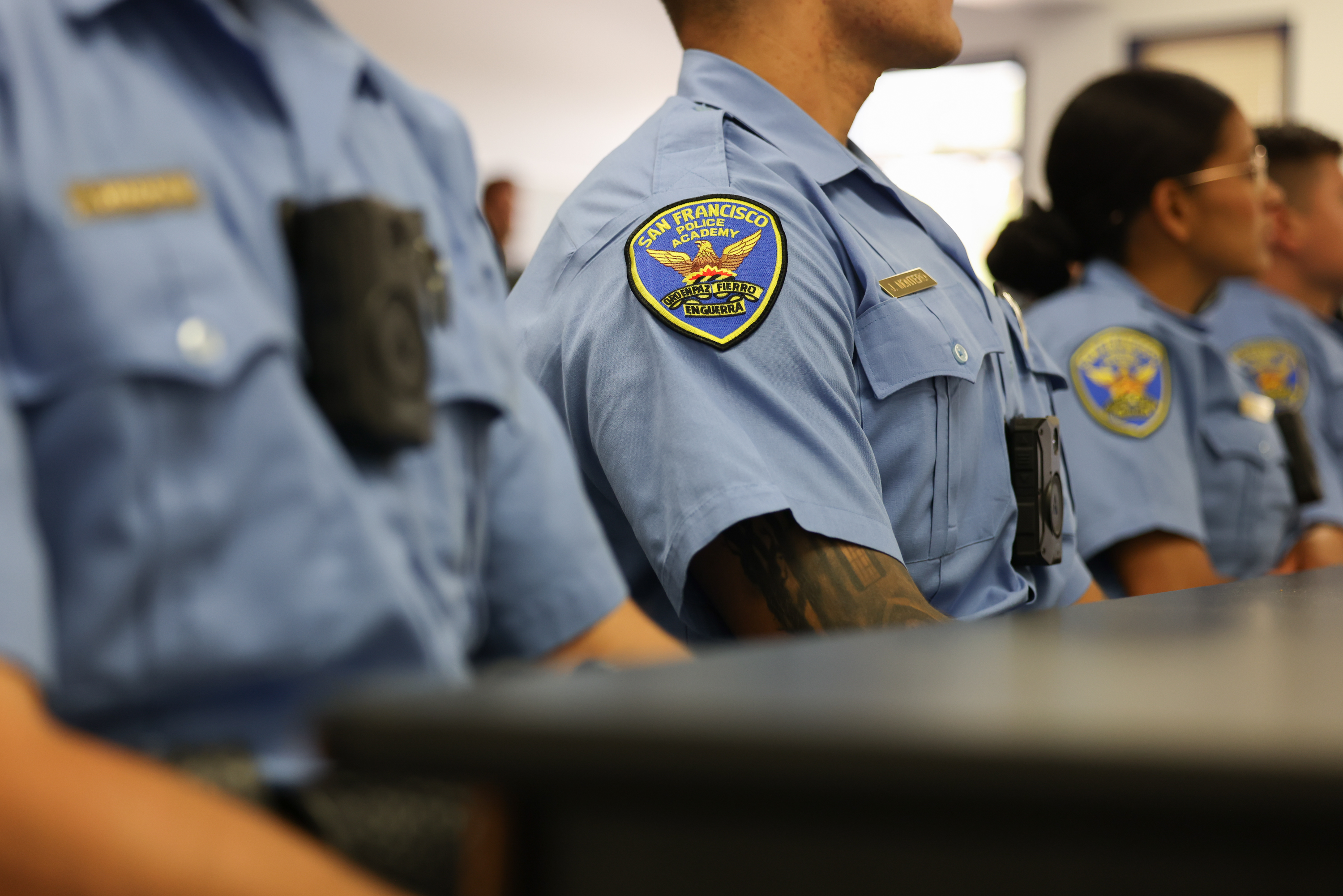 The image shows several police officers in light blue uniforms, sitting in a row. They wear body cameras and have &quot;San Francisco Police Academy&quot; patches on their sleeves.