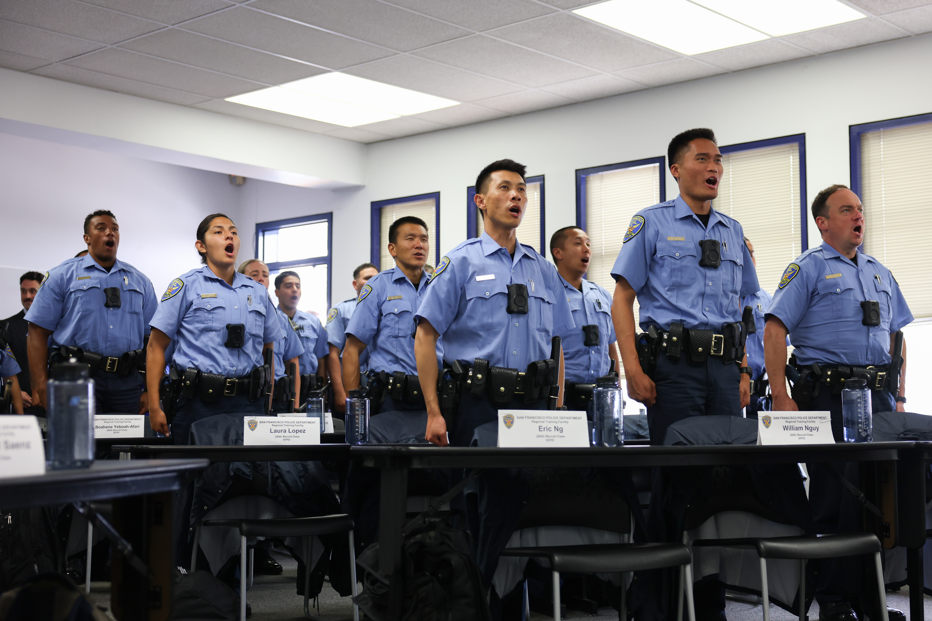 A group of uniformed police officers stands at attention, singing in unison in a classroom setting. Nameplates and water bottles are on desks in front.