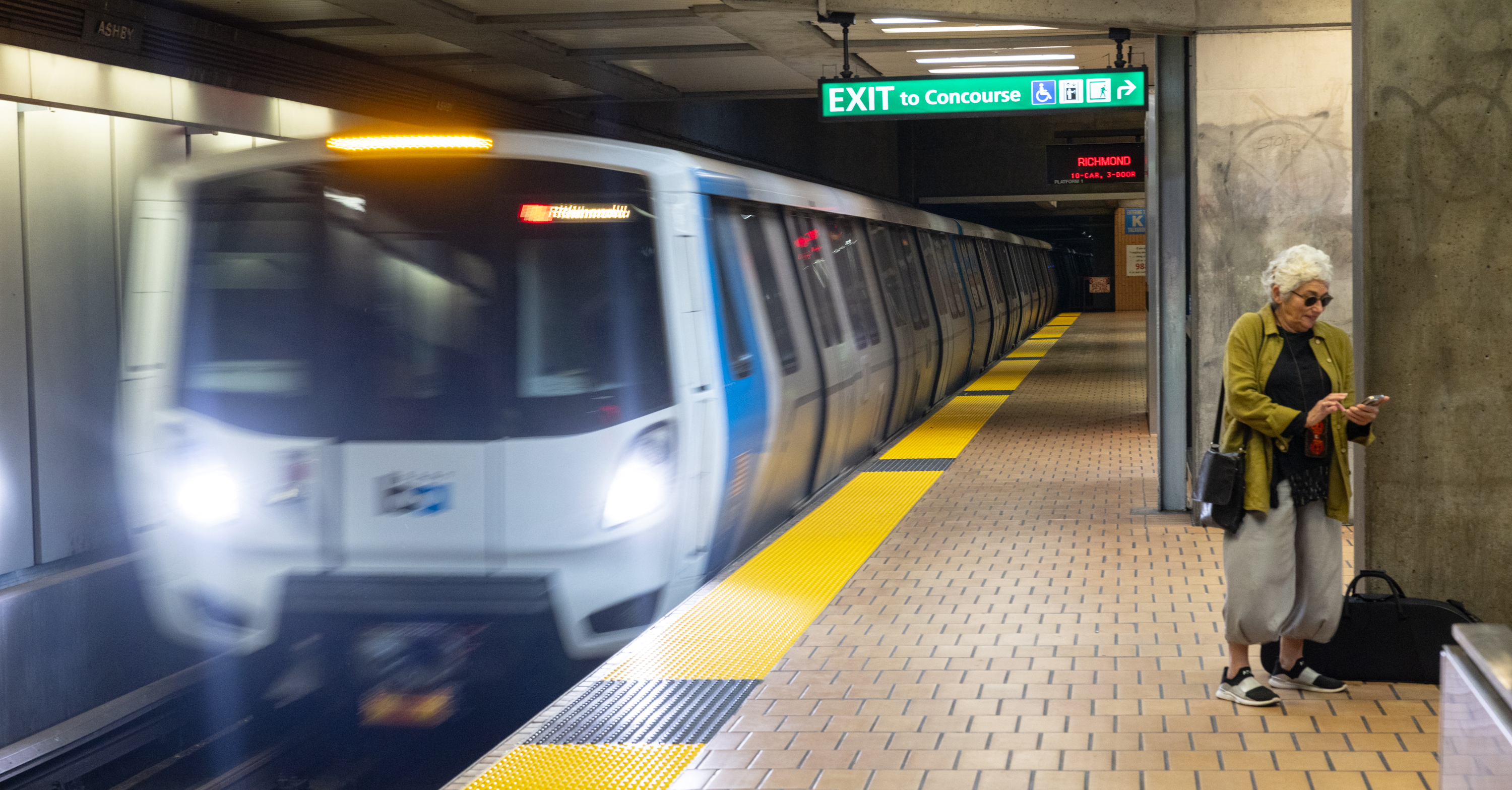 A subway train approaches the Ashby station as an elderly woman, wearing sunglasses and a green jacket, checks her phone beside a black suitcase near an "Exit to Concourse" sign.