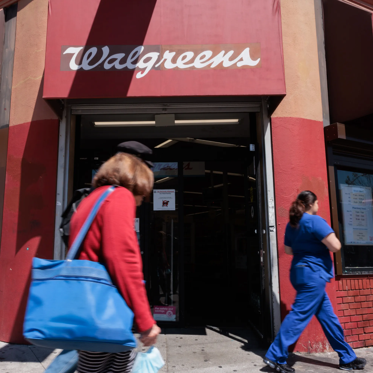 A storefront with a Walgreens sign, featuring a brick facade. Two people are walking by, one in a red coat holding a blue bag, and the other in blue scrubs.