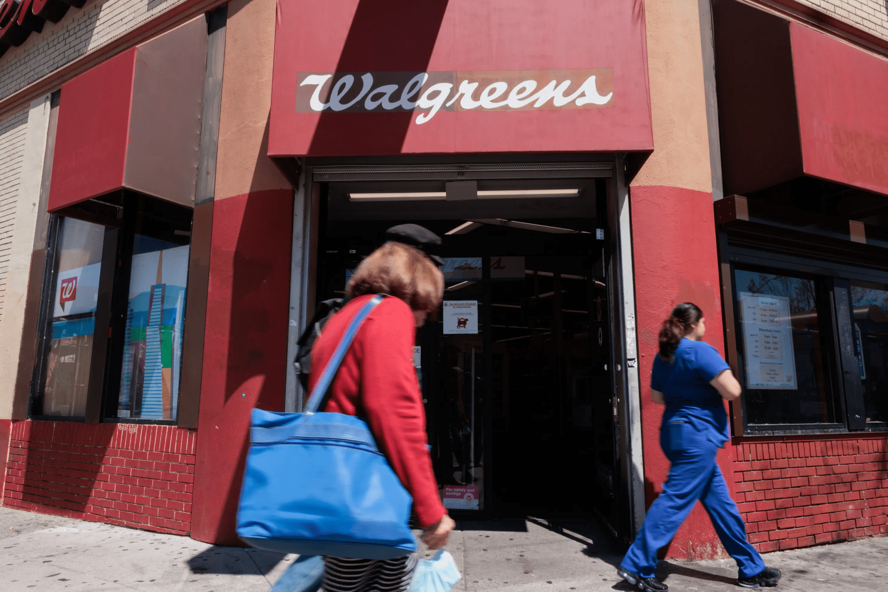 A storefront with a Walgreens sign, featuring a brick facade. Two people are walking by, one in a red coat holding a blue bag, and the other in blue scrubs.