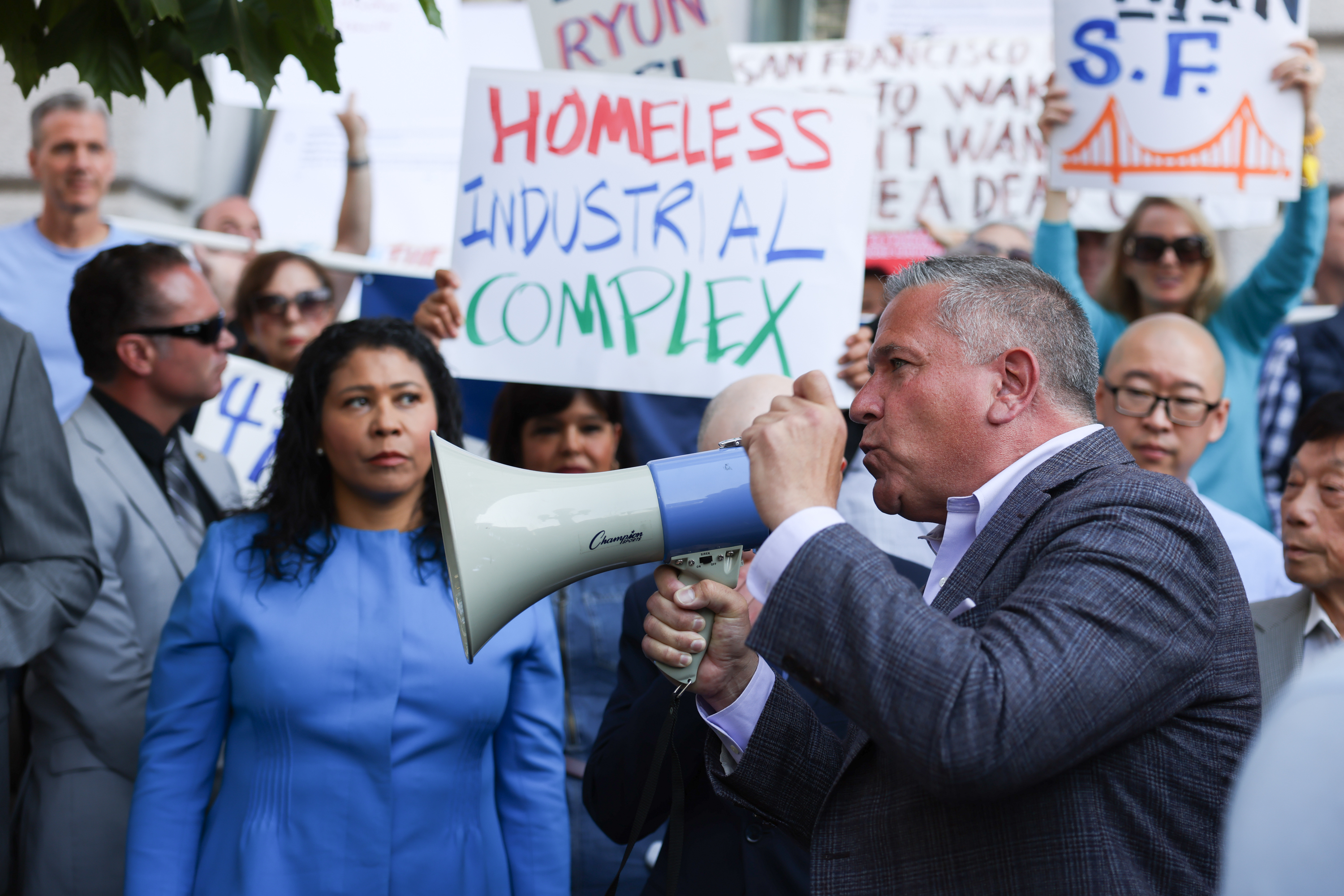 A man in a suit shouts into a bullhorn as someone holds a sign reading "homeless industrial complex" in the background.
