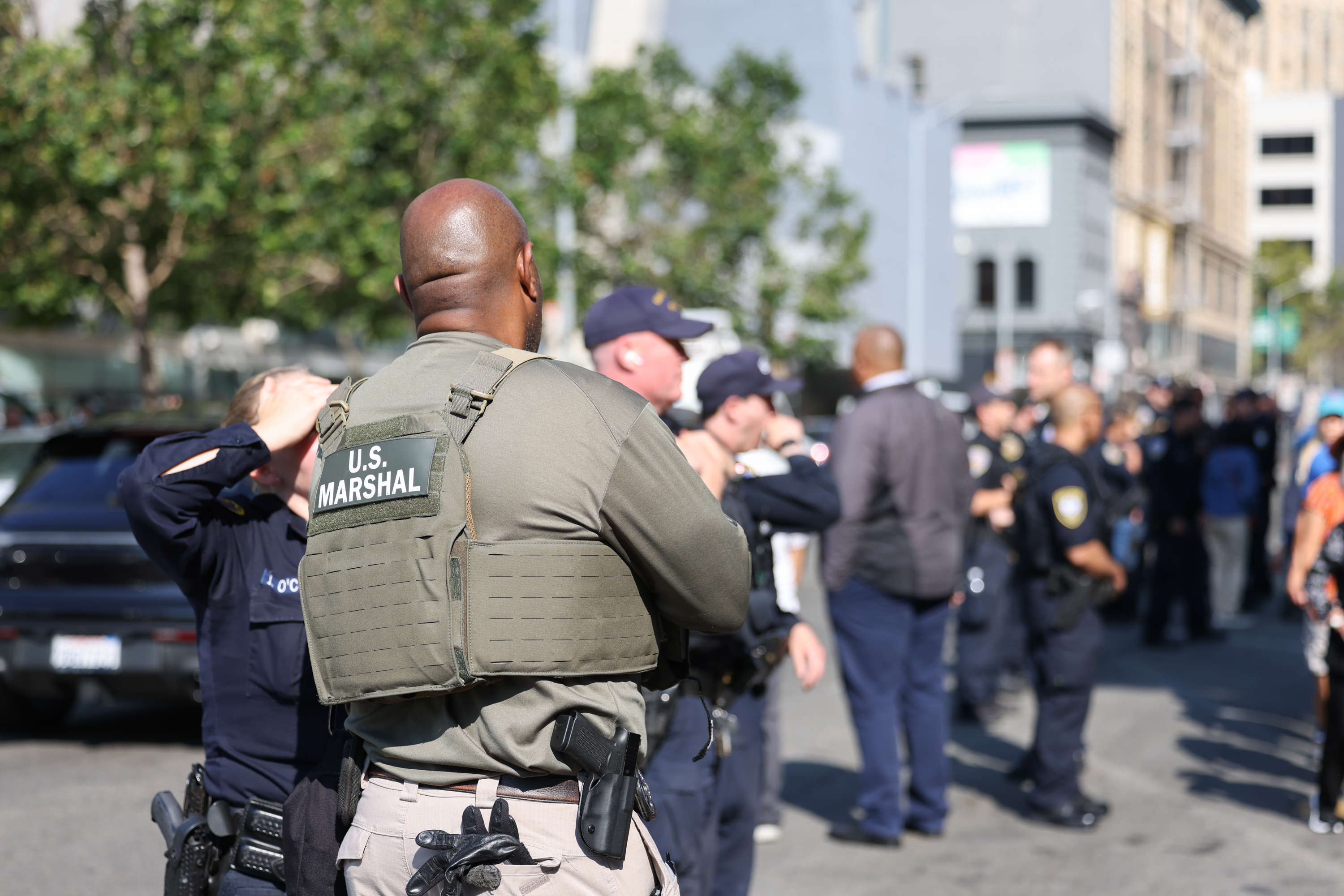 A group of law enforcement officers, including a U.S. Marshal, stand on a city street. The scene appears organized, with blurred buildings and trees in the background.