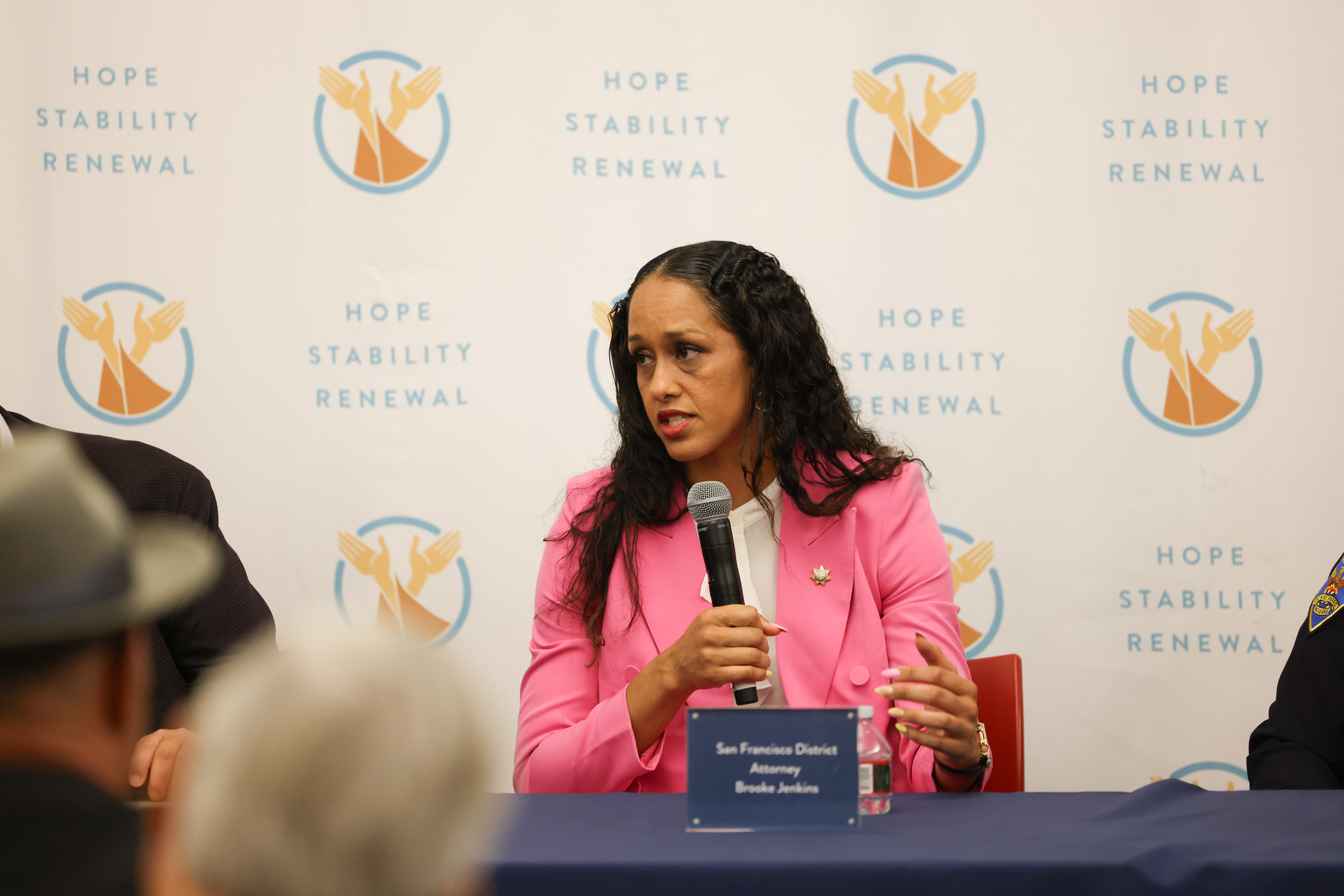 A woman in a pink jacket speaks into a microphone at a table with a backdrop displaying &quot;Hope Stability Renewal&quot; logos.