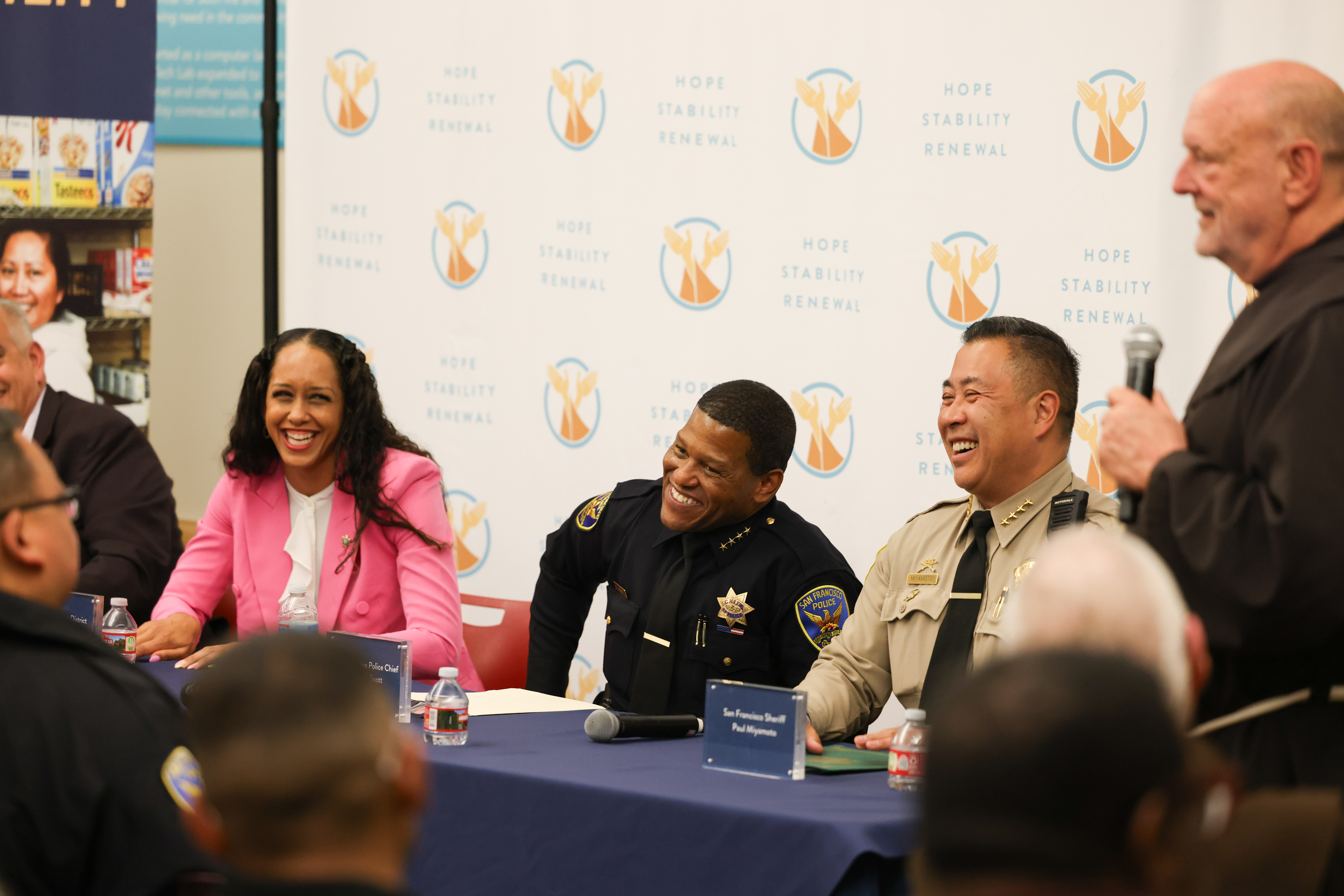 Several people are seated at a table, smiling and laughing. They are in uniform and business attire, with a backdrop labeled &quot;Hope Stability Renewal.&quot;