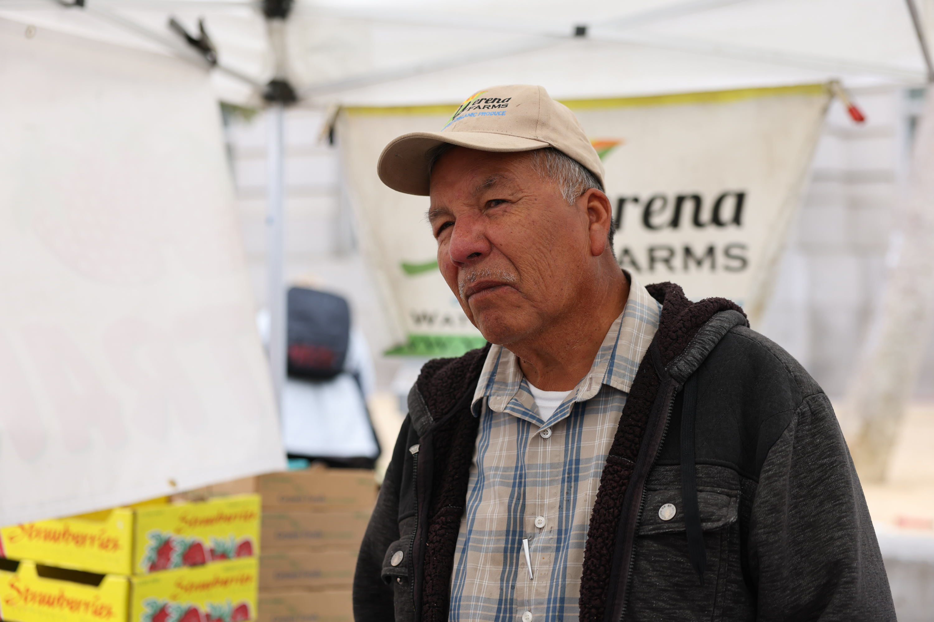 An older man wearing a cap and jacket stands in front of a tent with farm labels.
