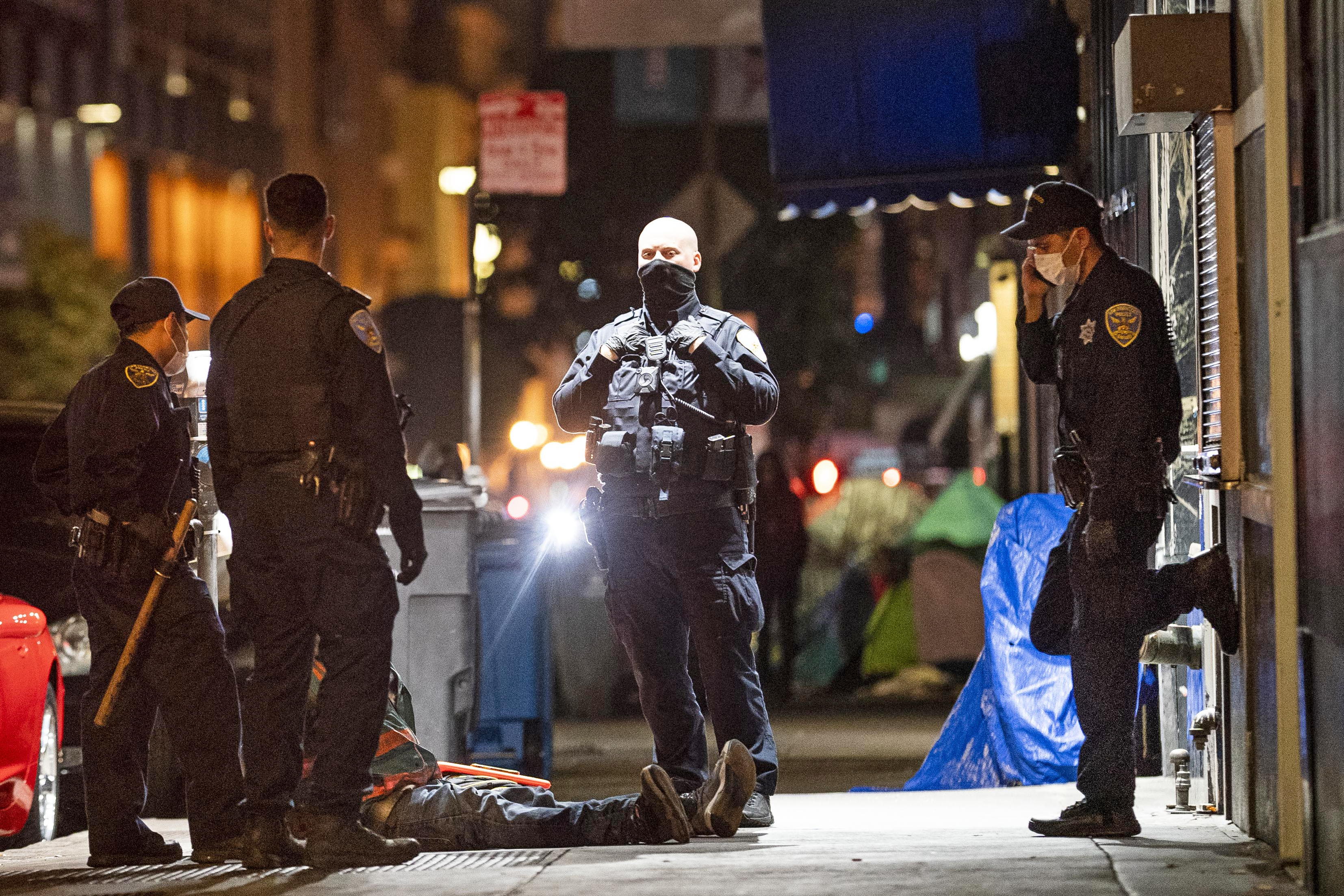 Police officers at a nighttime crime scene with a body covered by a tarp on the sidewalk.