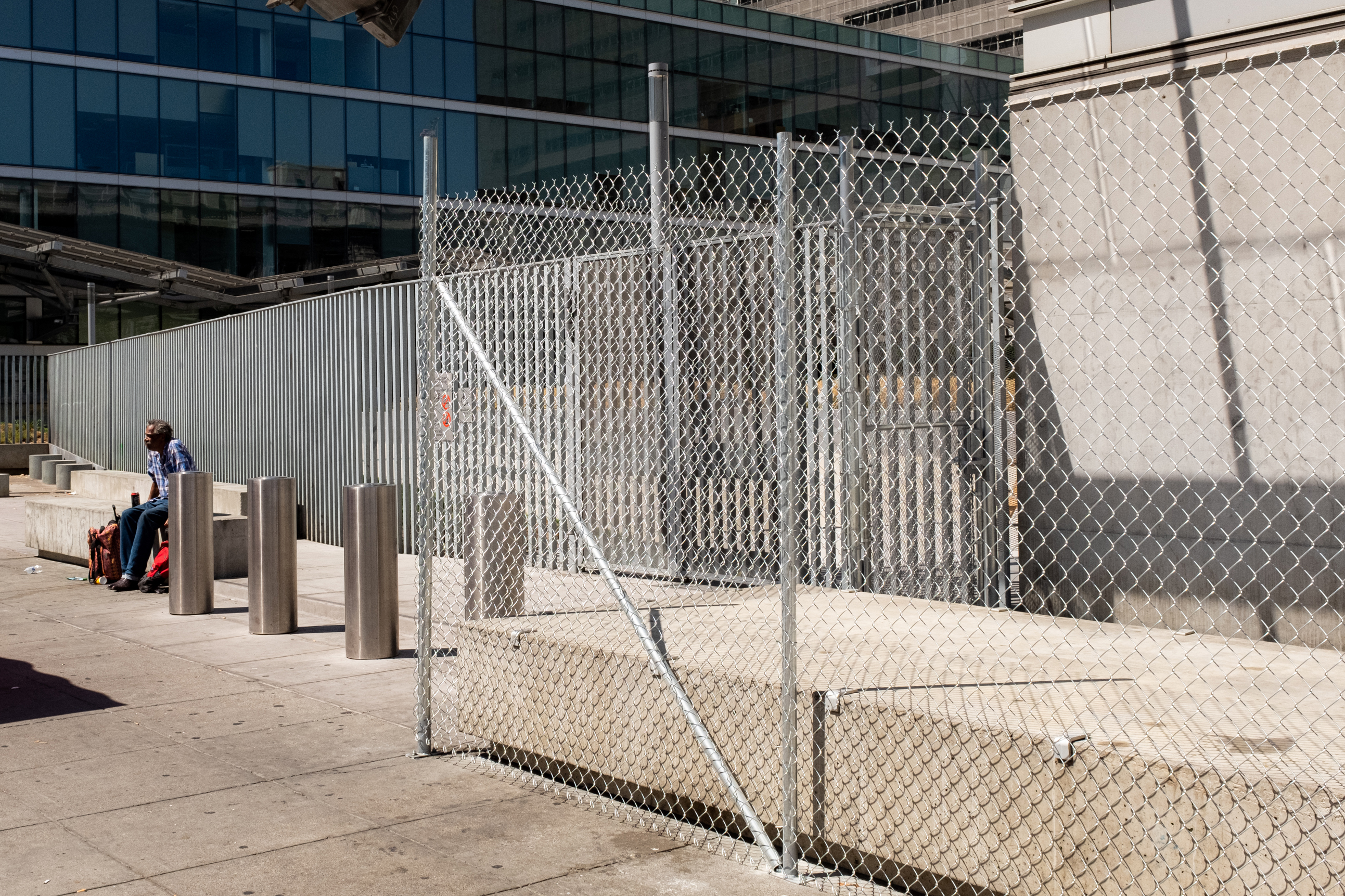 A man sits by metal bollards on a concrete sidewalk next to a chain-link fence. Behind is a modern building with large glass windows.