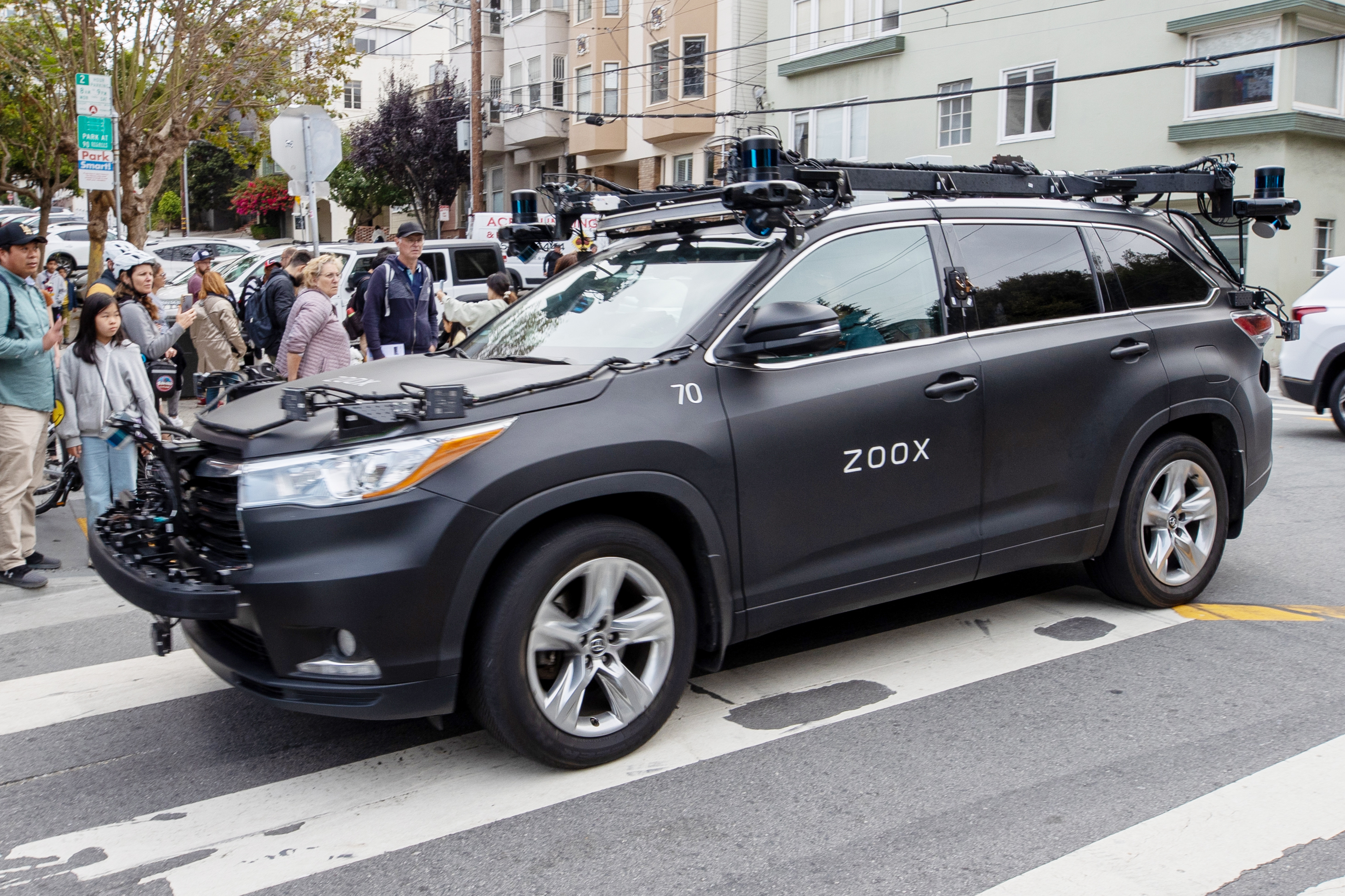 A black SUV with a Zoox logo on the left front door drives on Lombard Street as pedestrians watch.