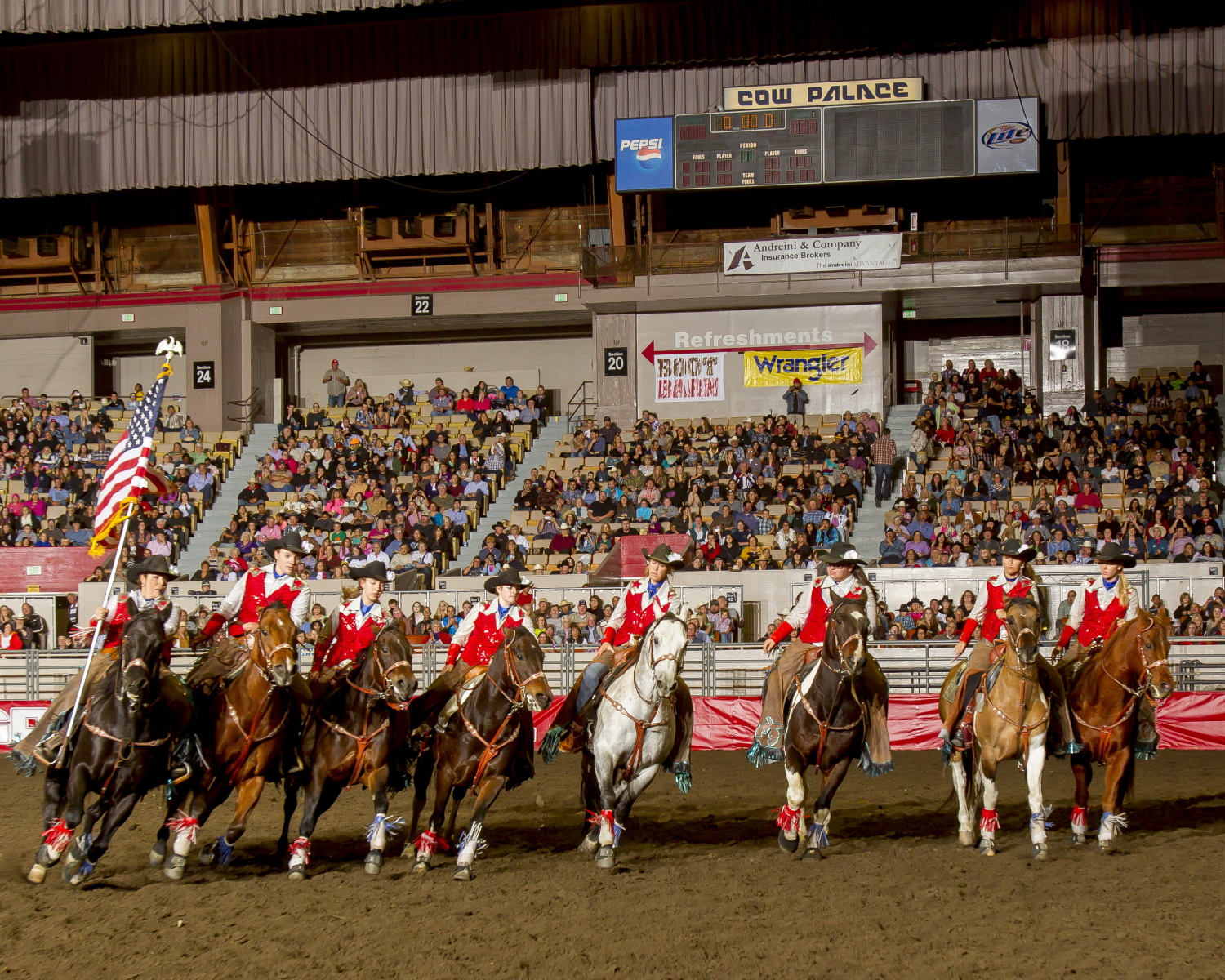 A team of riders on horses carrying flags parades in an arena at the &quot;Cow Palace&quot; in front of a large audience.
