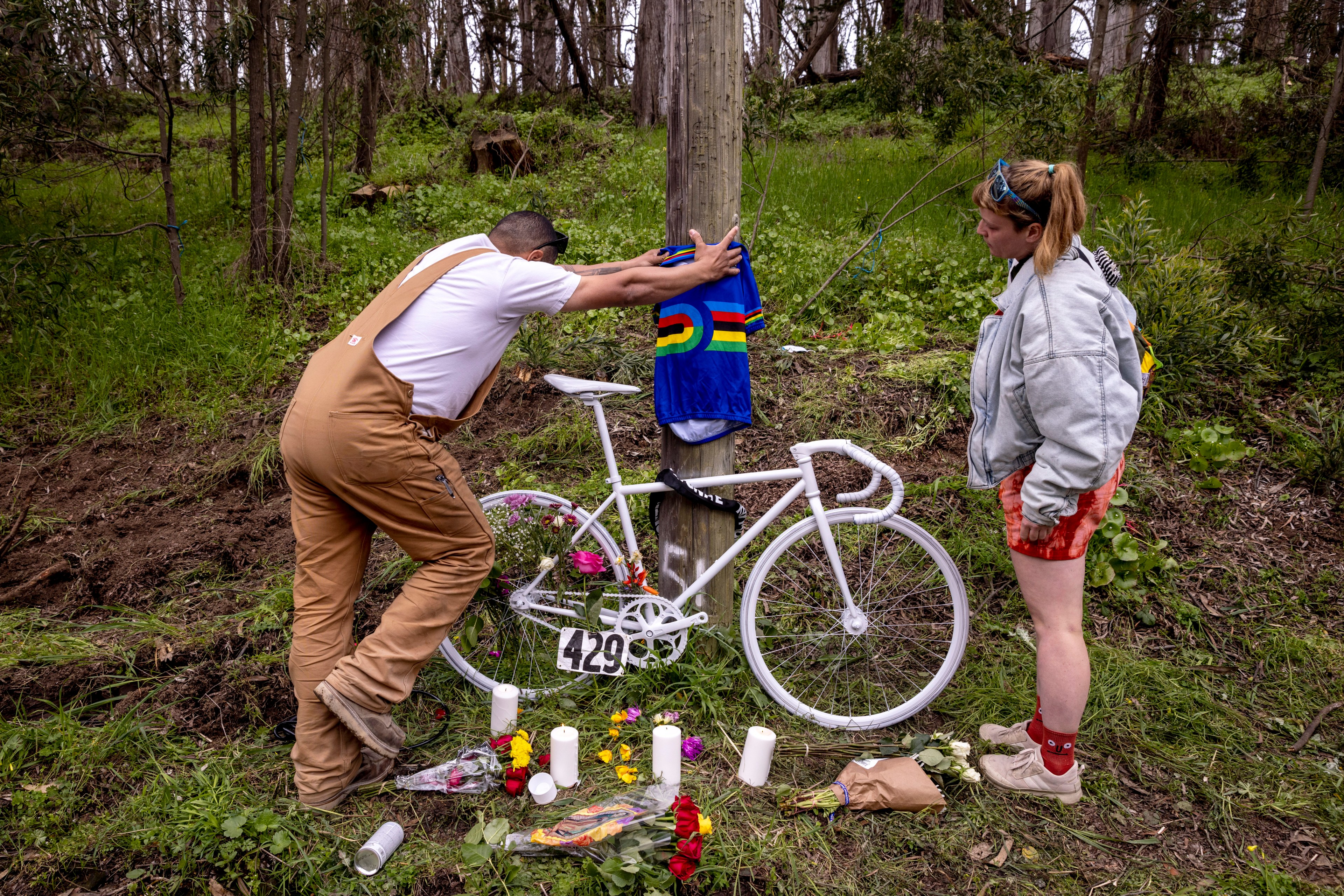 Two people stand beside a white-painted bike at a roadside memorial adorned with flowers.
