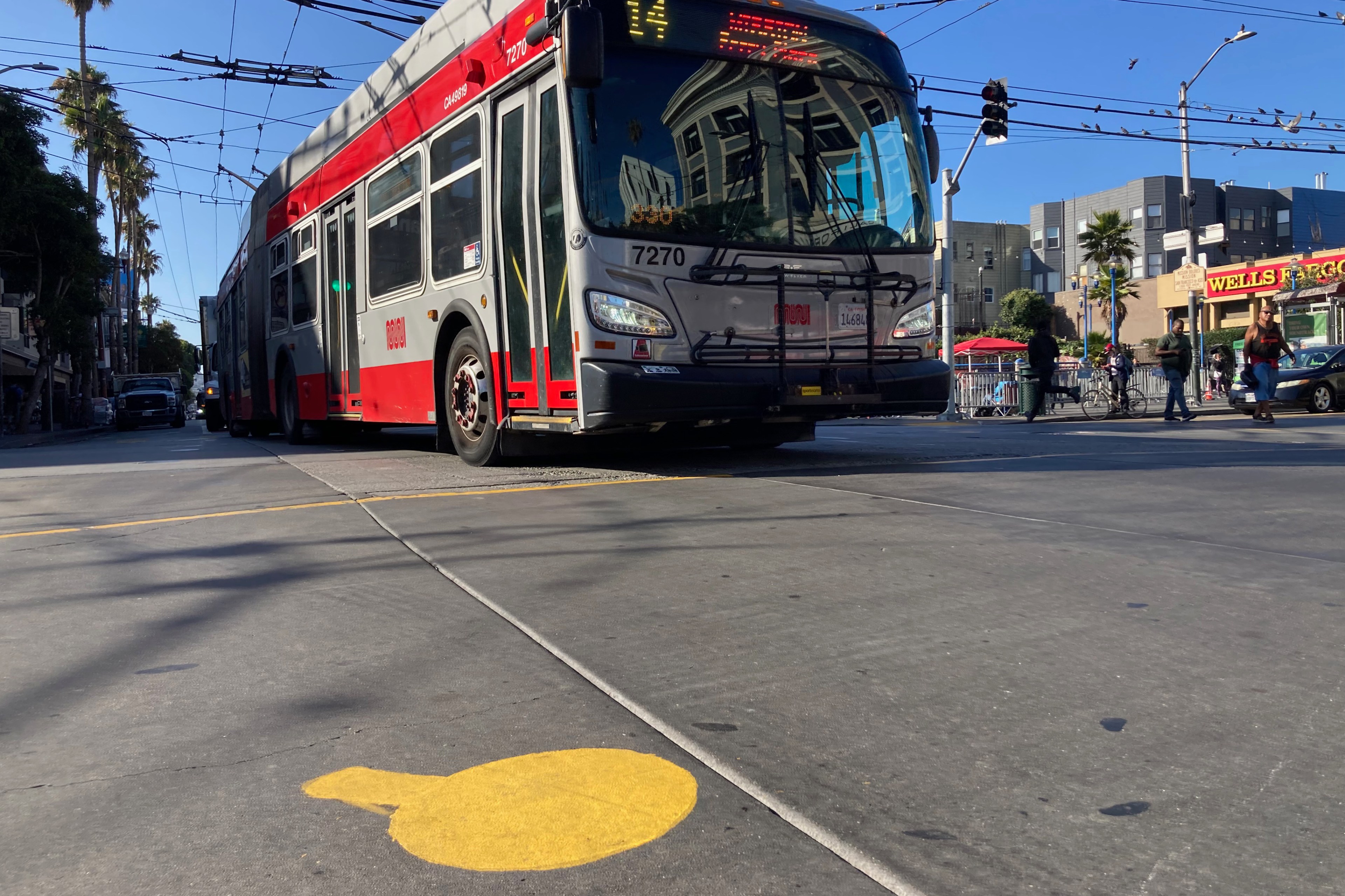 A red and silver articulated bus is turning on a city street with overhead wires, near pedestrians and shops.