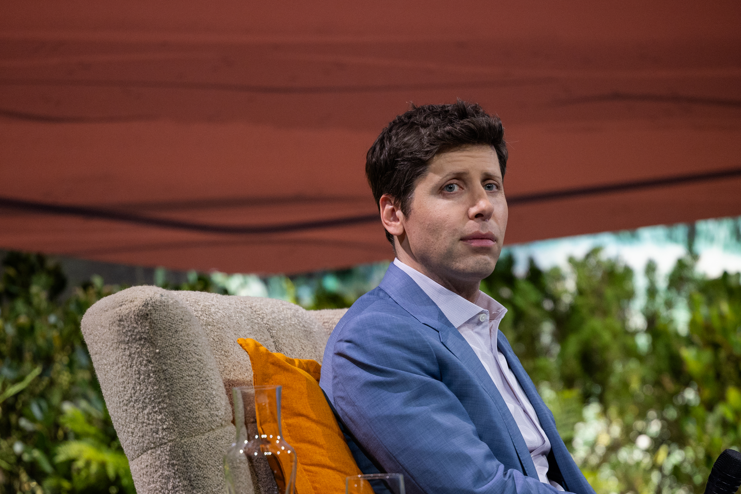 A man in a blue suit and white shirt is sitting on a beige chair with an orange cushion. In the background, there are green plants and a reddish-brown canopy.