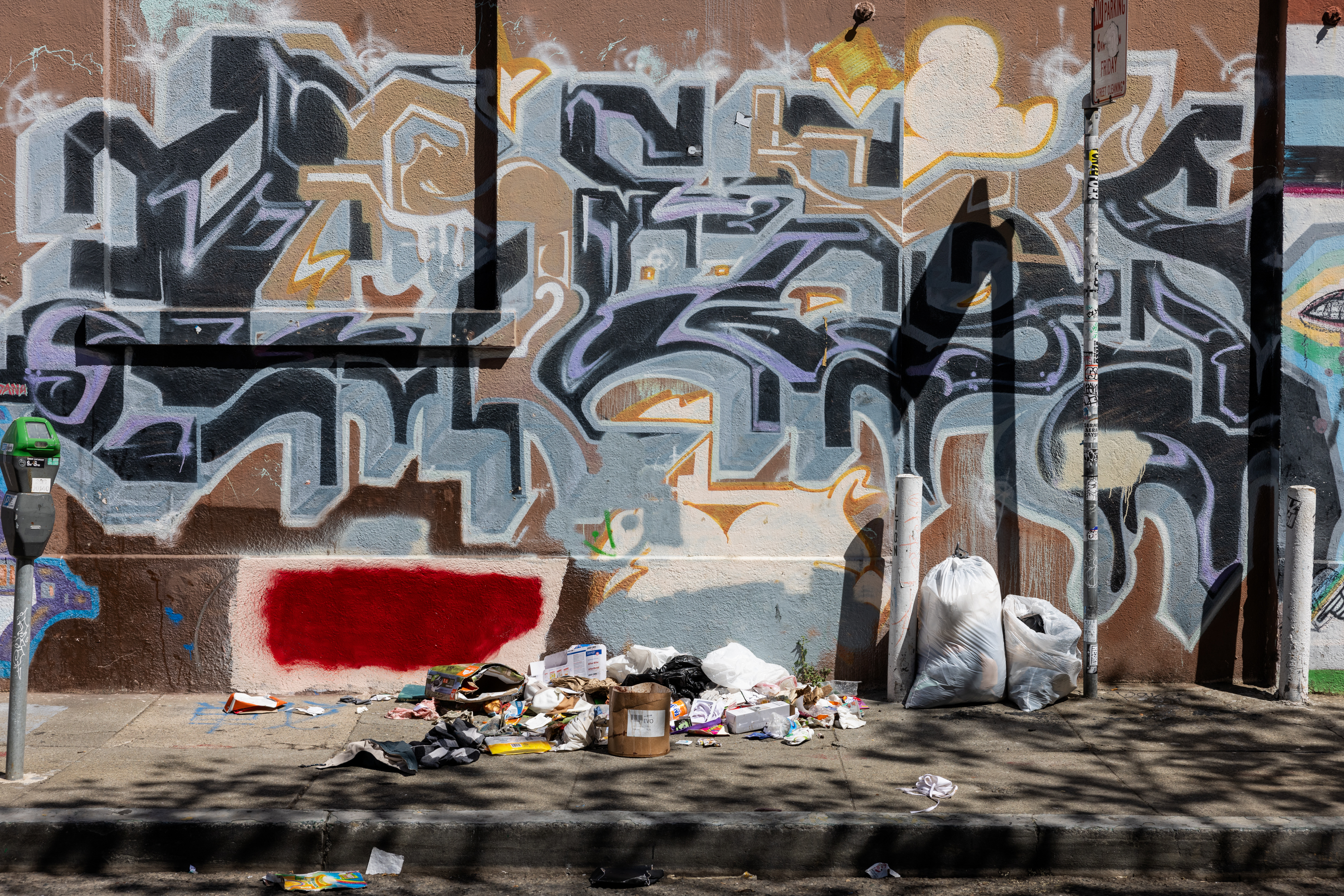 A wall with colorful graffiti art is in the background, while trash and bags of garbage are scattered on the sidewalk in front. A parking meter is also visible.