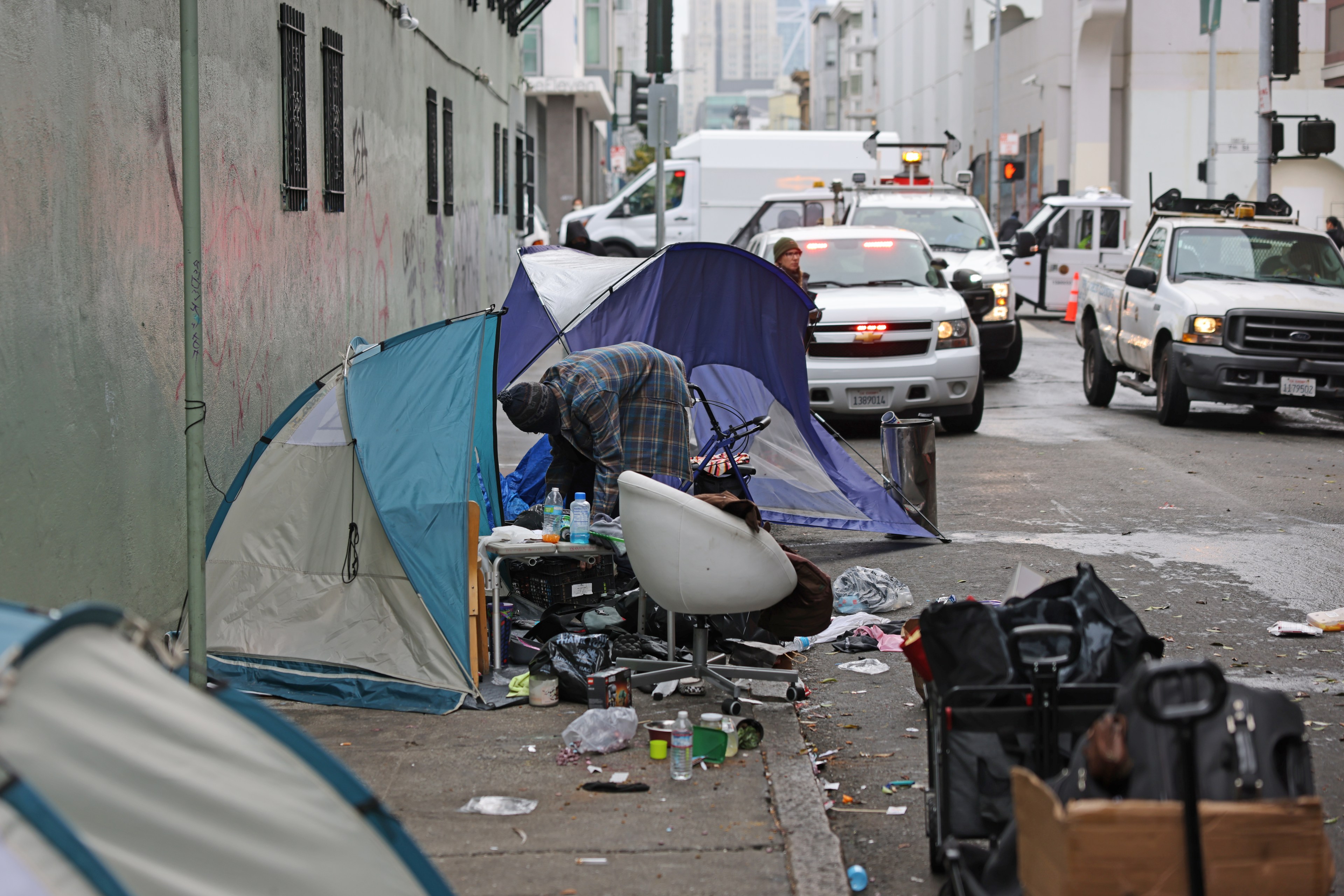 An alley with tents, a person rummaging through belongings, scattered trash, and cars in line.