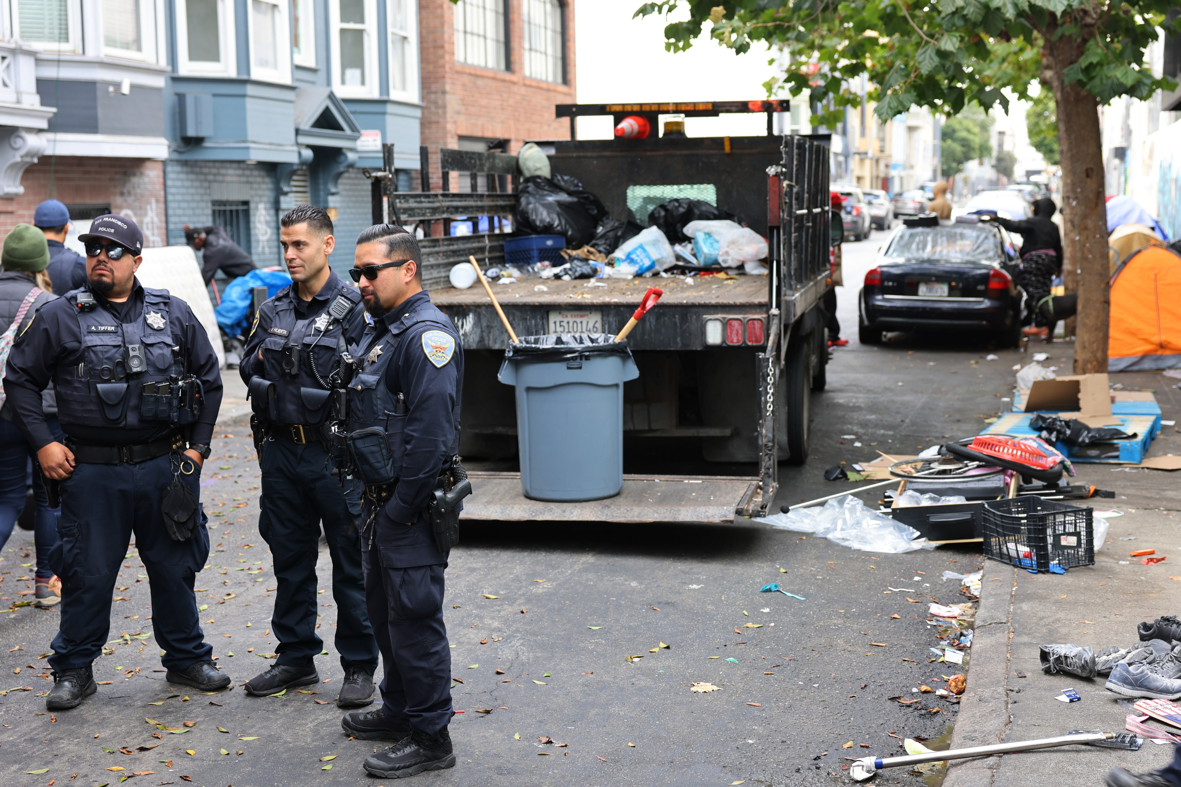 Three officers stand by a garbage truck and litter on an urban street, with tents in the background suggesting a homeless encampment.