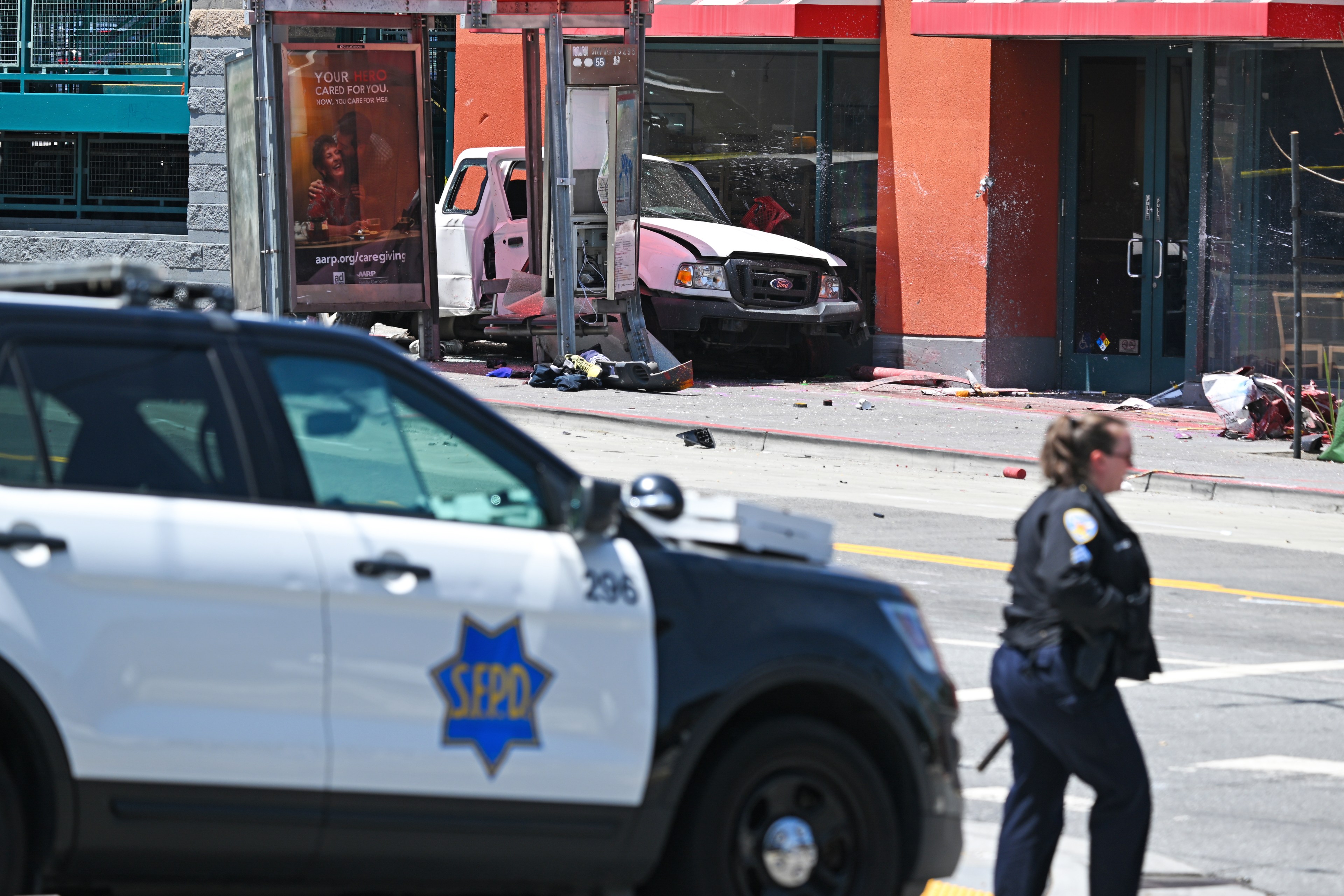 In the background, a white Ford truck is crashed into a bus stop and a retail building. In the foreground, a police officer, who has just exited her vehicle, approaches on foot.