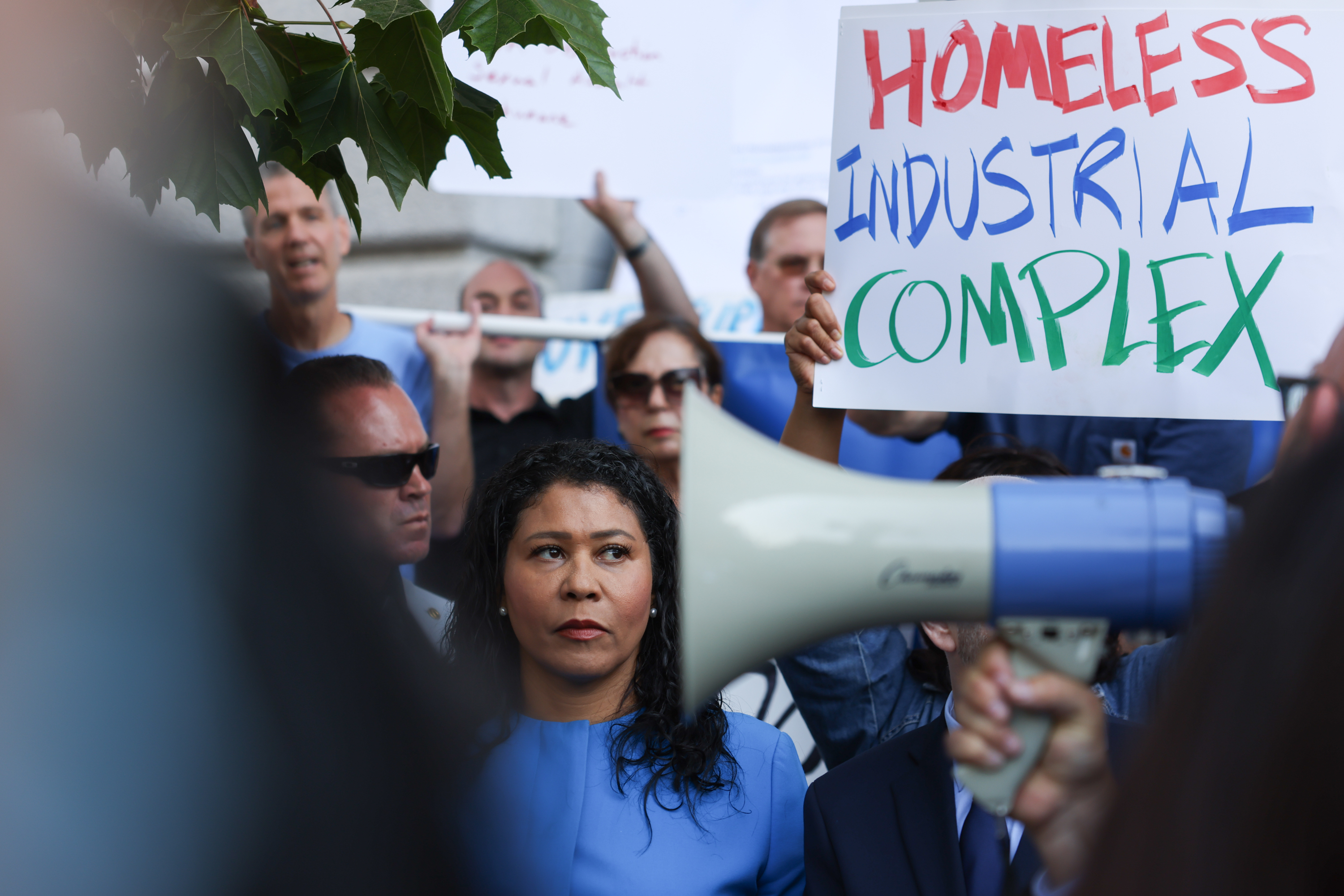 A woman with a megaphone is in focus with protesters and a "HOMELESS INDUSTRIAL COMPLEX" sign in the background.