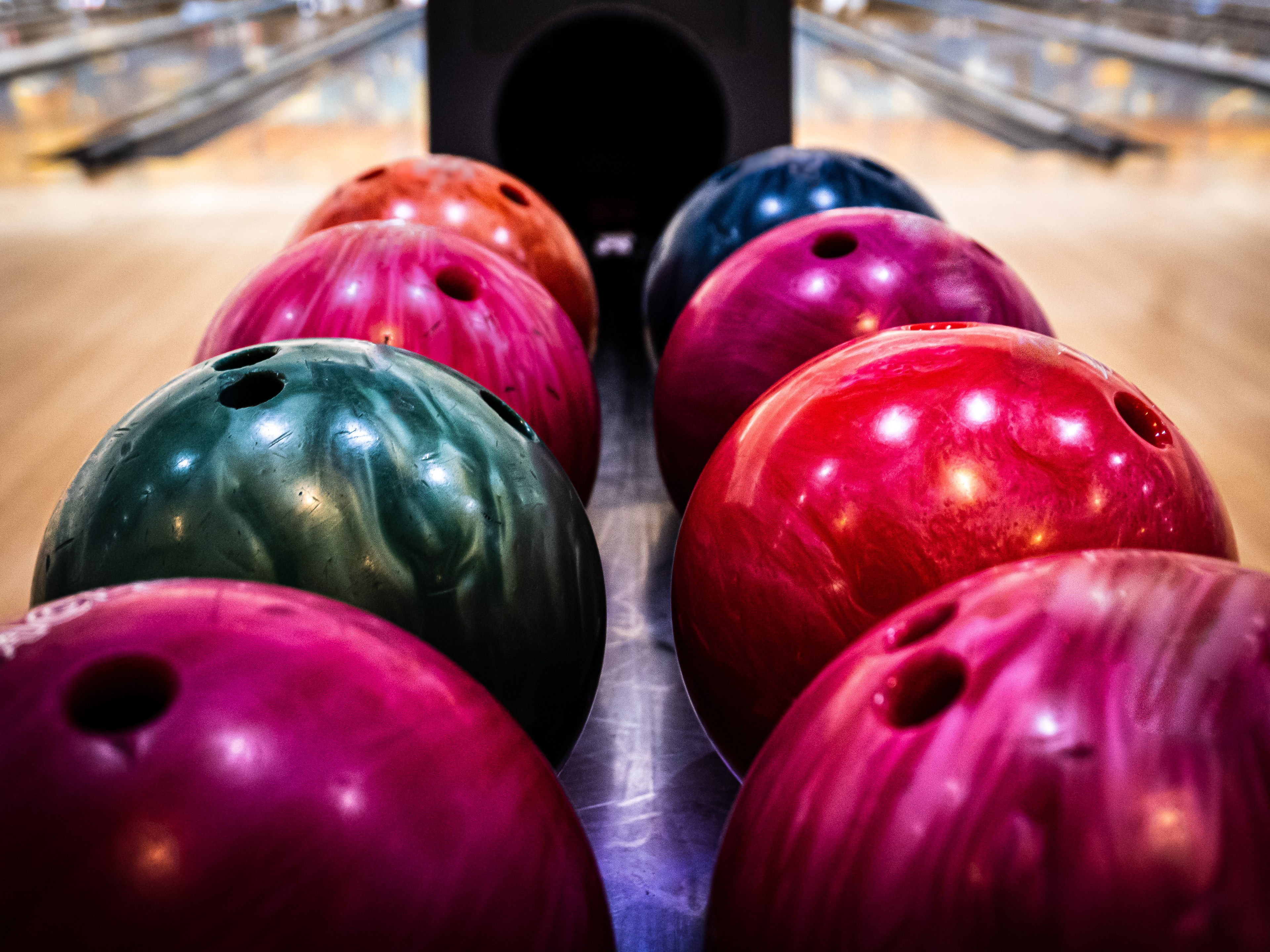Colorful bowling balls lined up on a rack, with a bowling lane visible in the background.