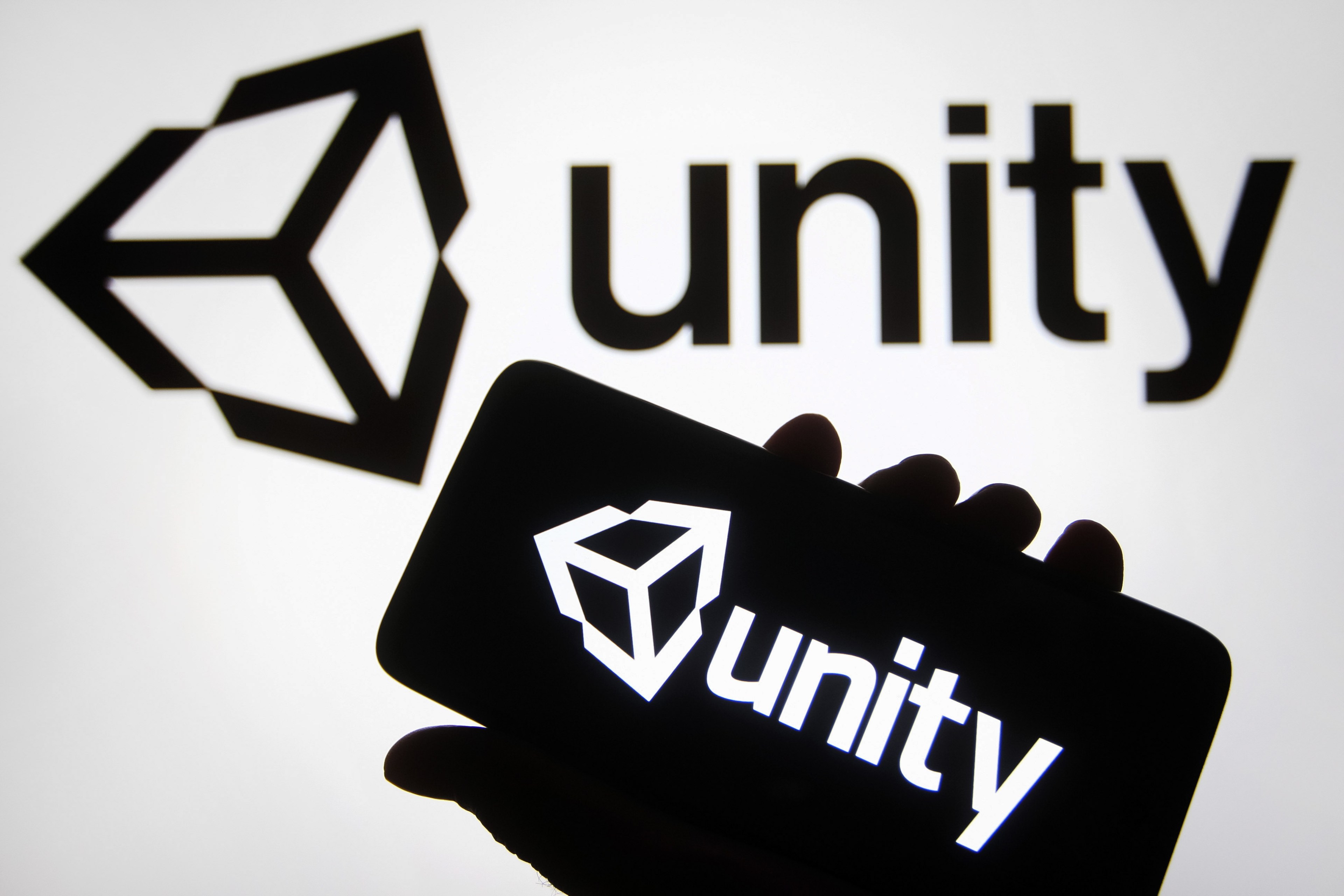 A hand is holding a smartphone with the Unity logo on the screen. Behind the hand is a computer screen with the logo.