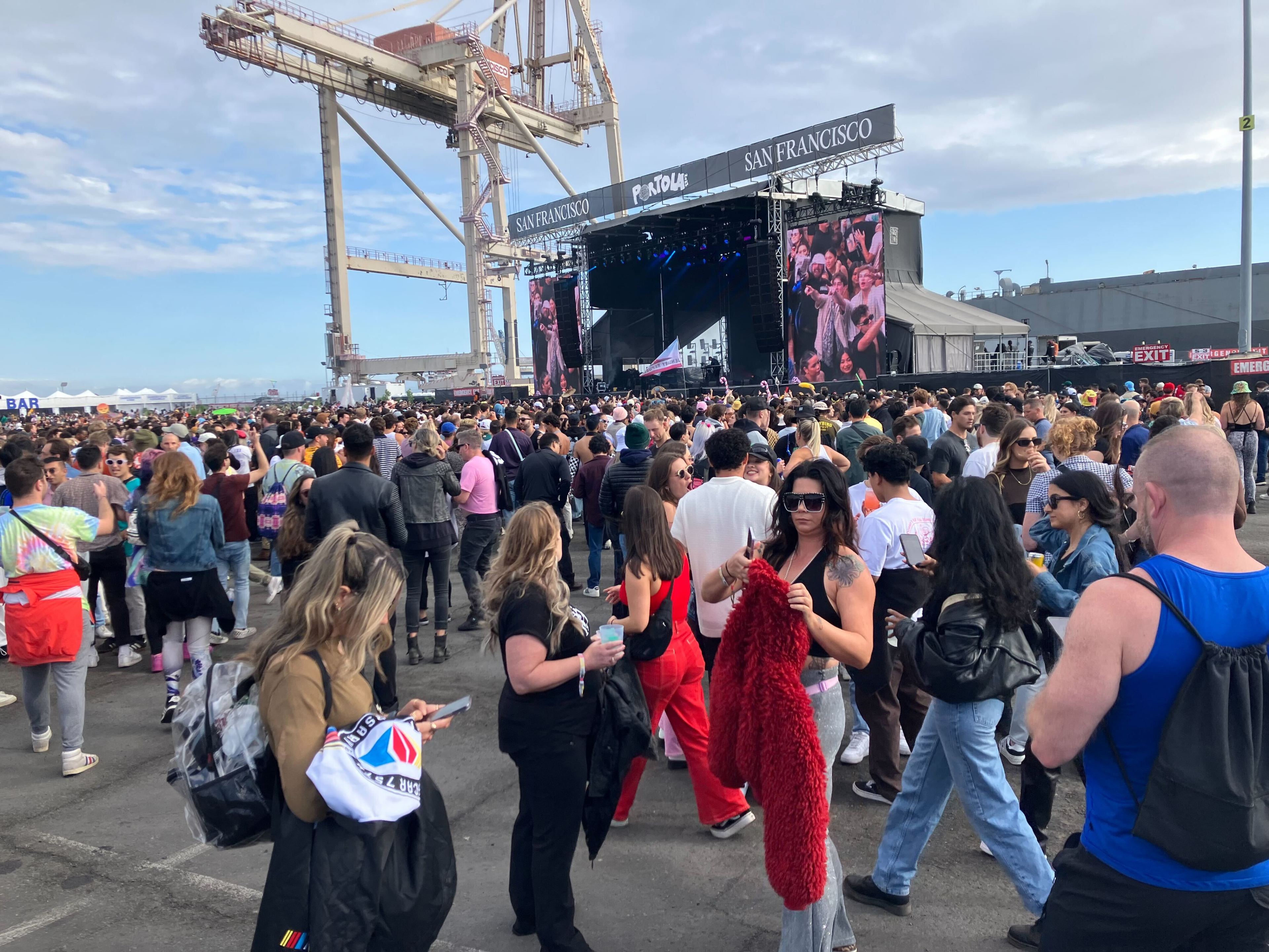A lively outdoor music festival crowd near a stage with large screens, under daylight with industrial cranes in the background.