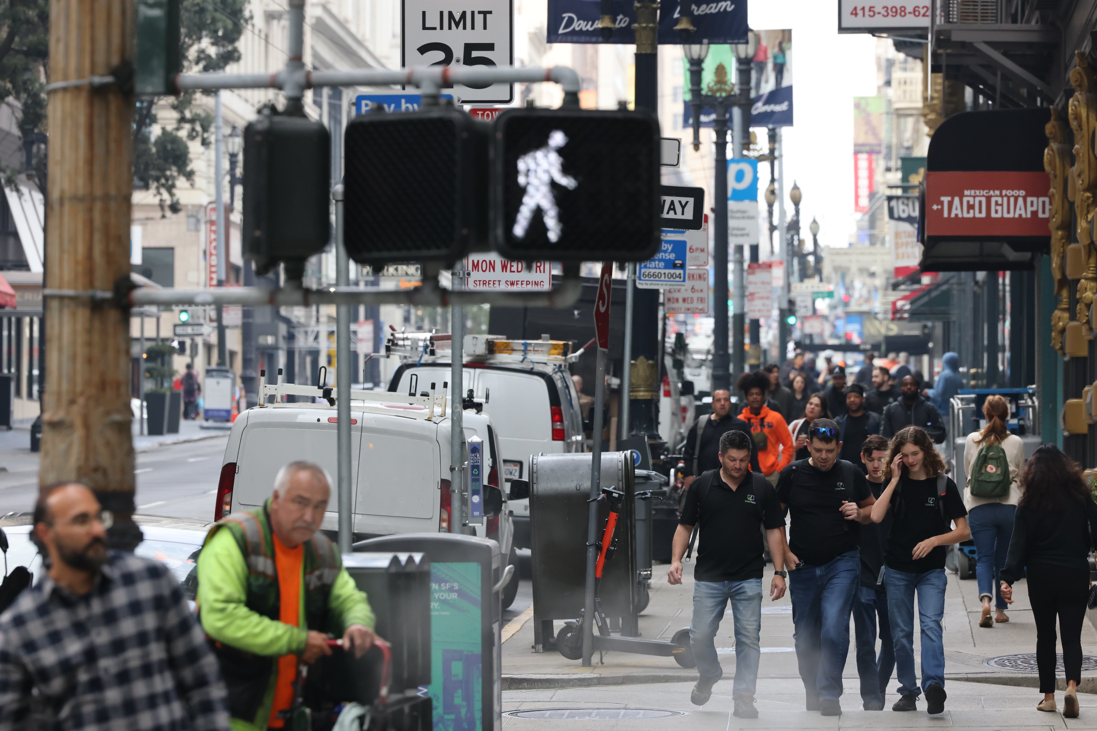 A bustling city street scene with pedestrians walking on the sidewalk. Traffic signs include a walking signal and speed limit. Vans and storefronts line the street.