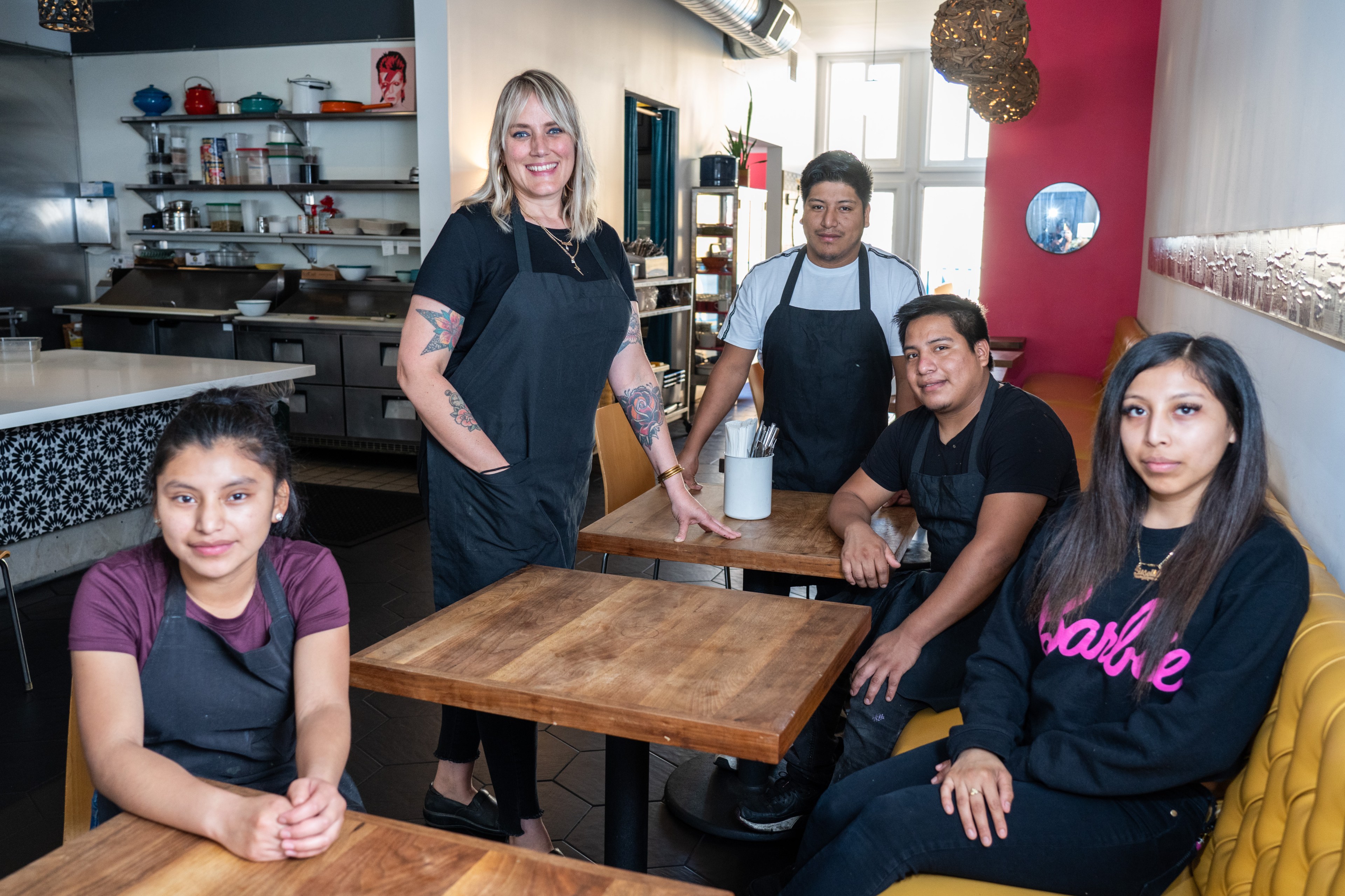 A diverse restaurant team poses inside a cozy eatery with a kitchen in the background.