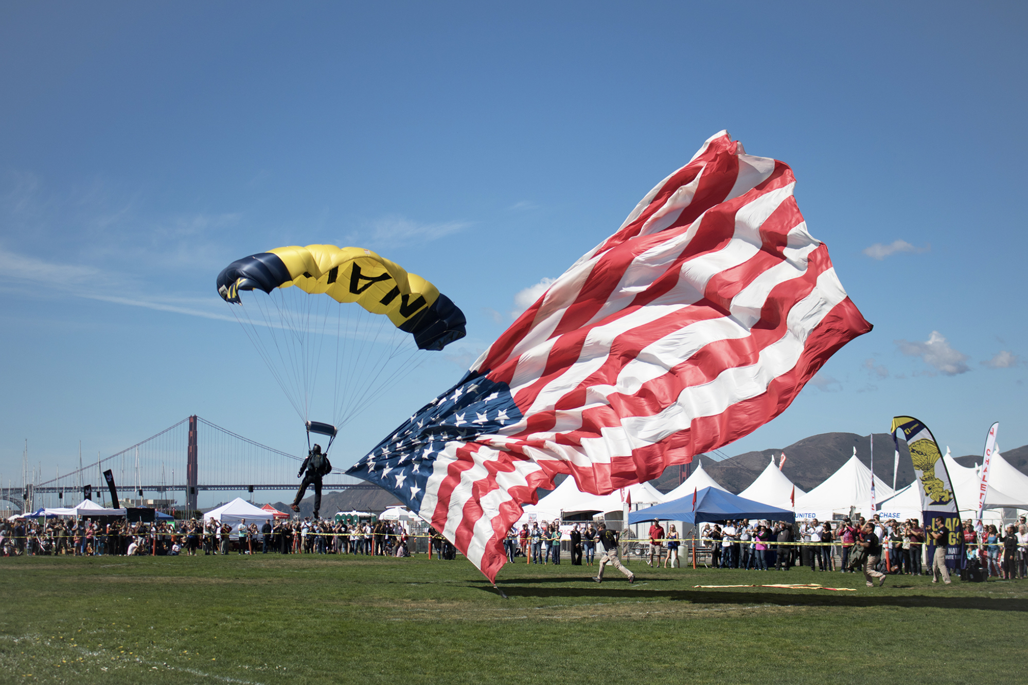 A parachutist lands smoothly while holding a large American flag, with an event crowd and the Golden Gate Bridge visible in the background on a clear day.