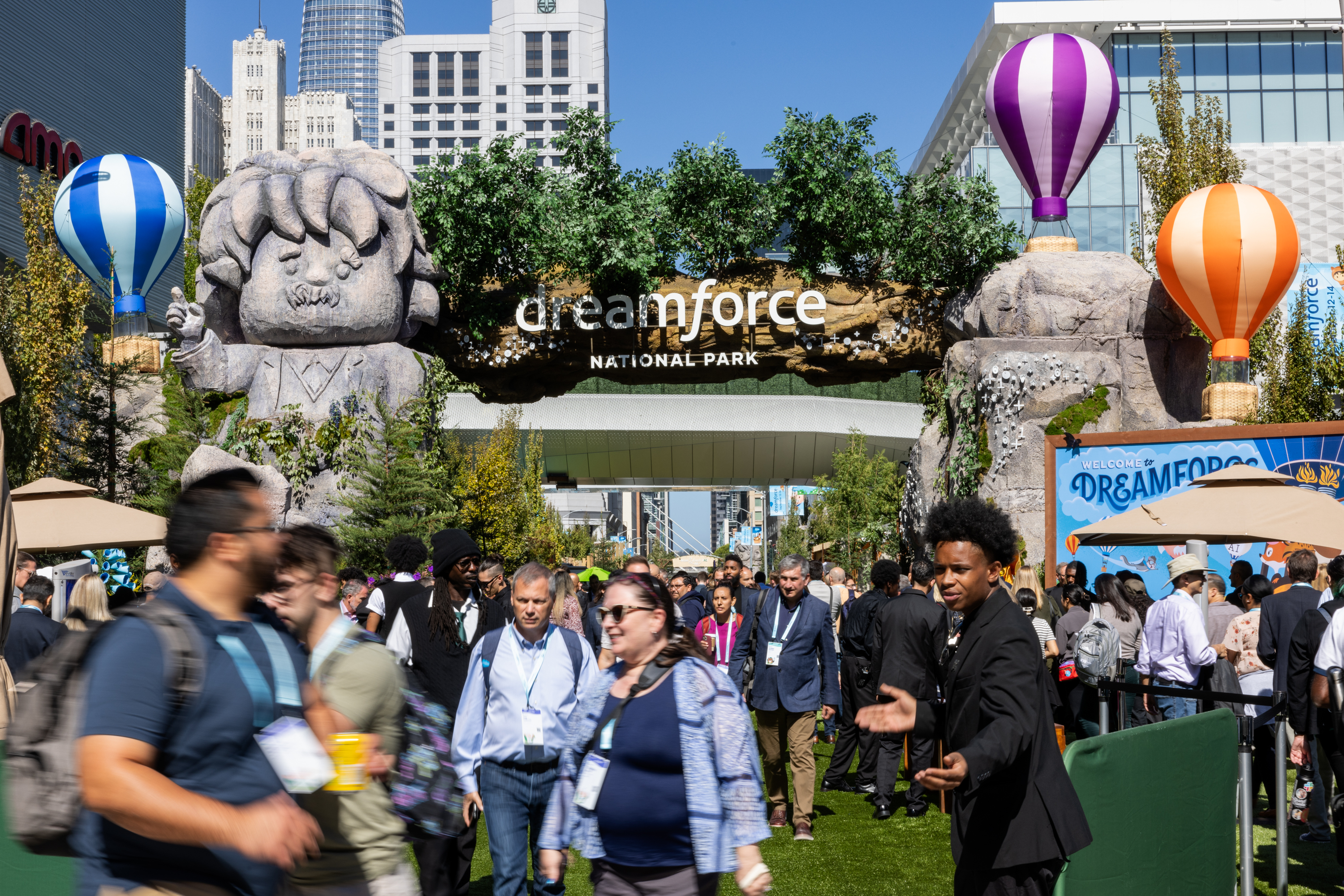 The image shows a crowded event entrance labeled &quot;Dreamforce National Park,&quot; decorated with large, cartoonish statues, colorful hot air balloons, and green foliage under a clear sky.