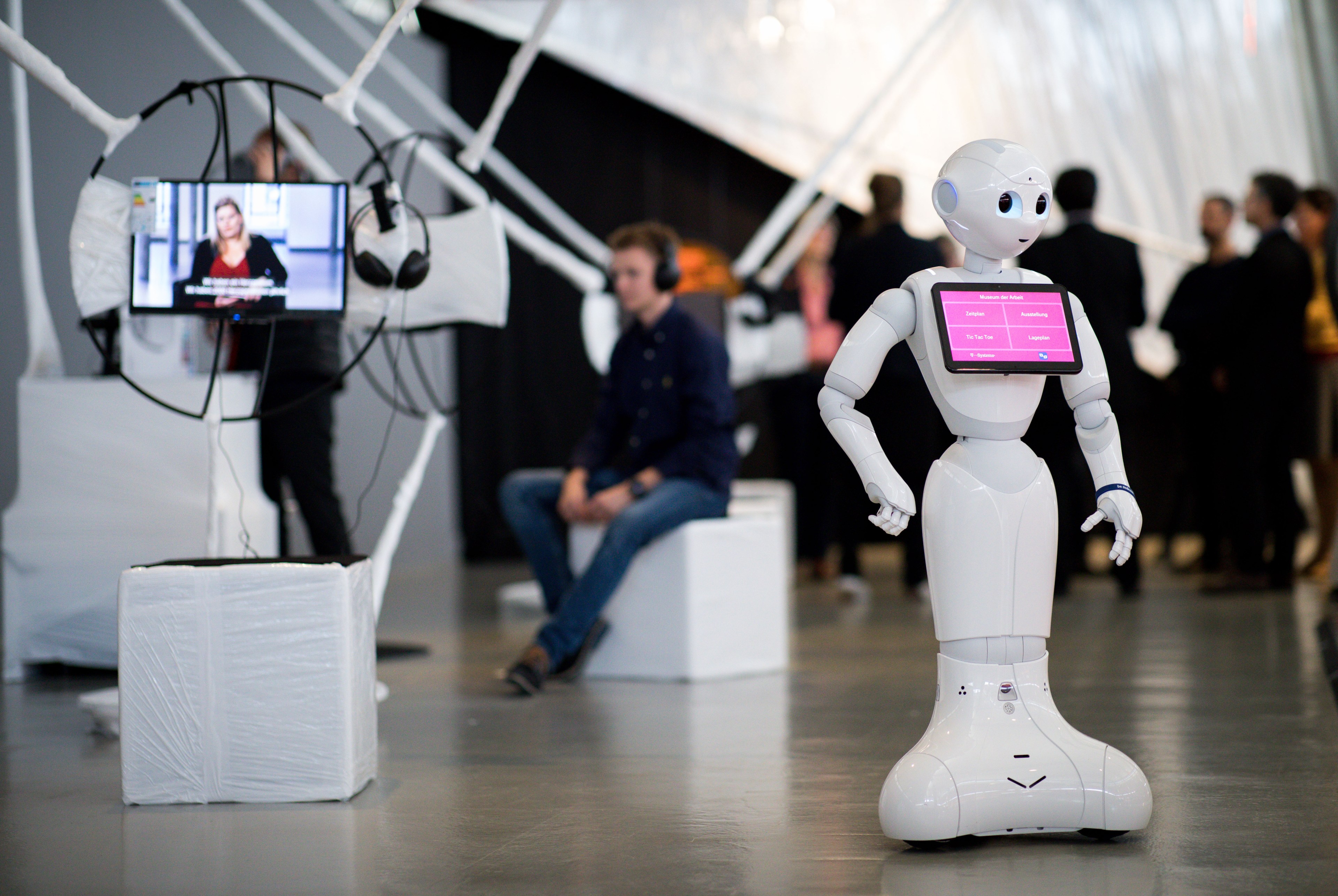 A humanoid robot with a screen on its chest stands in an exhibit hall near a man seated in the background.