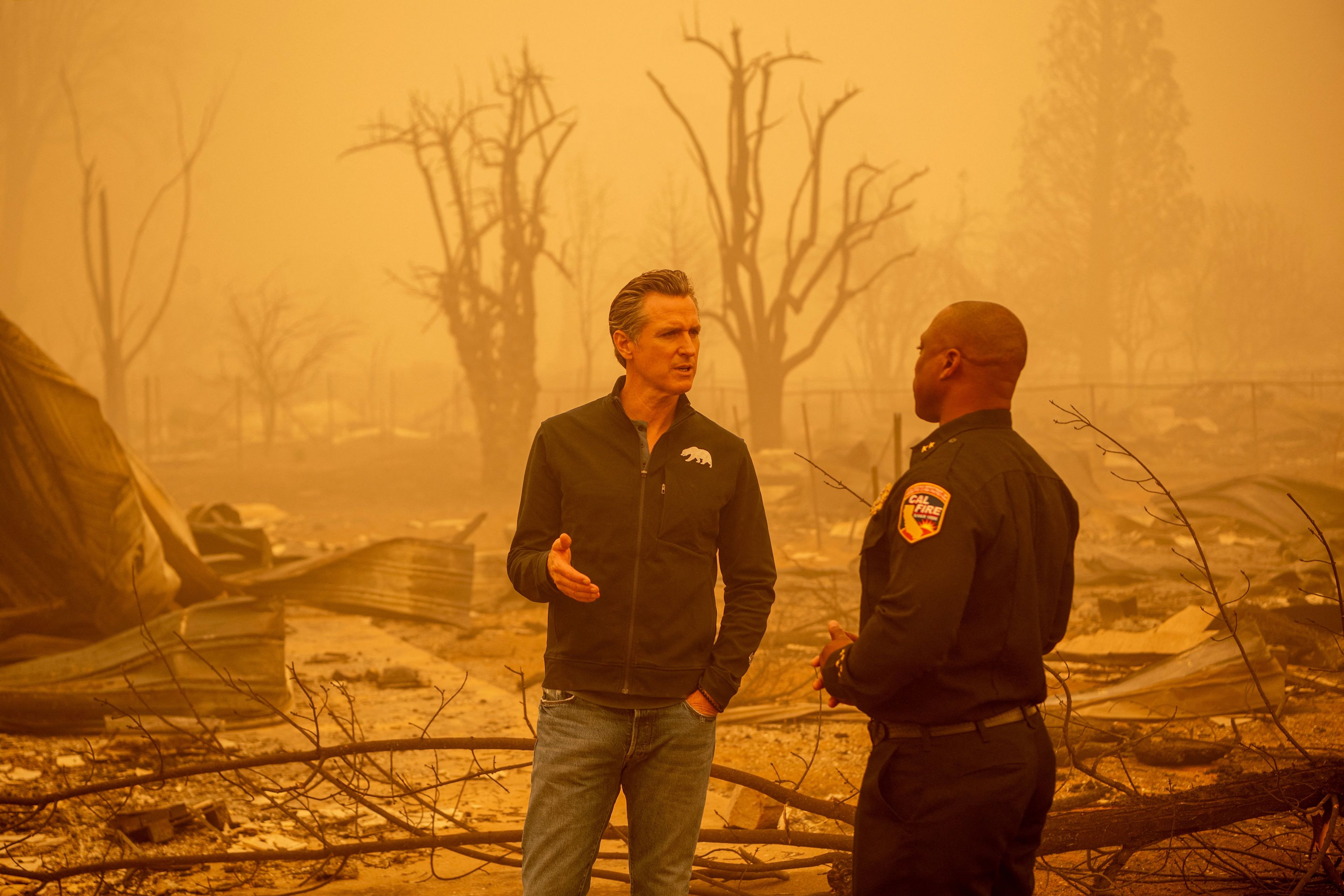 Two men, Gov. Gavin Newsom and a Cal Fire official, stand talking in front a burnt landscape surrounded by orange haze.