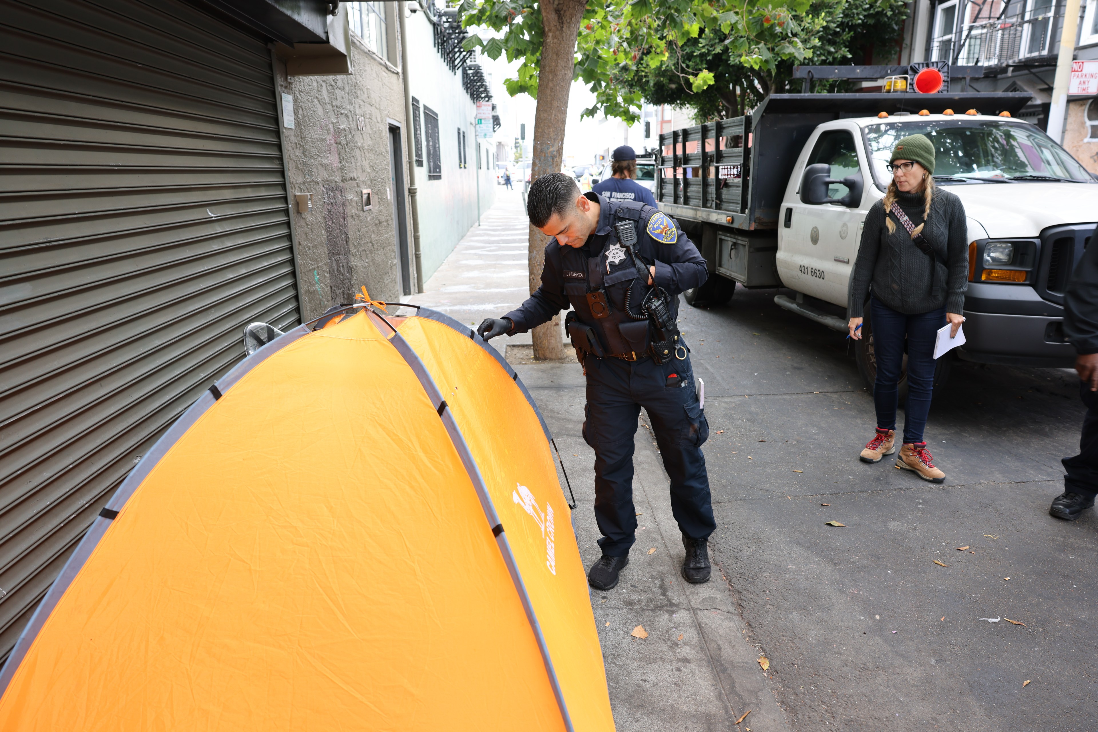 A police officer inspects an orange tent on a sidewalk; a woman and a vehicle are nearby.
