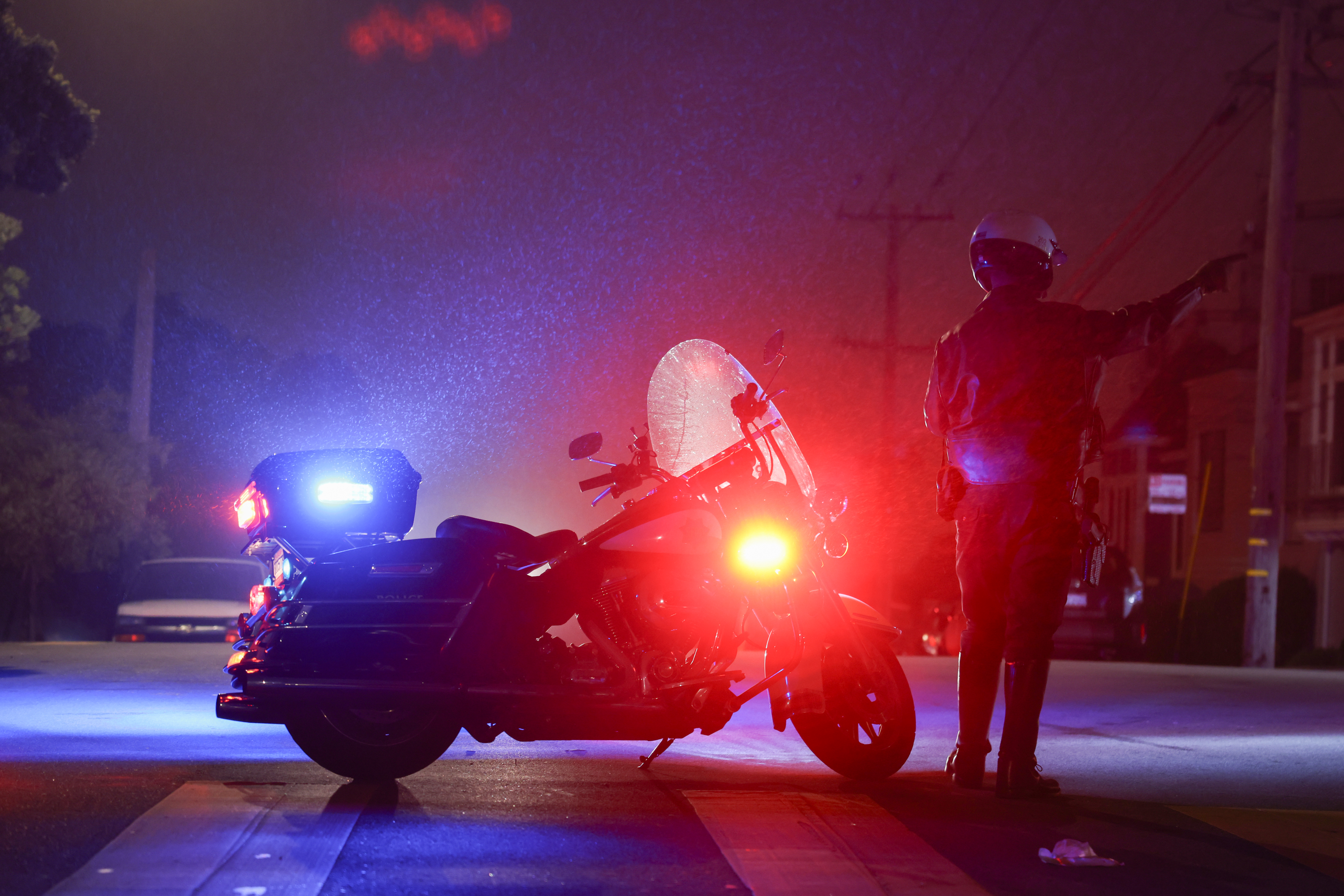 A police officer stands next to a motorcycle with flashing lights on a misty street at night.