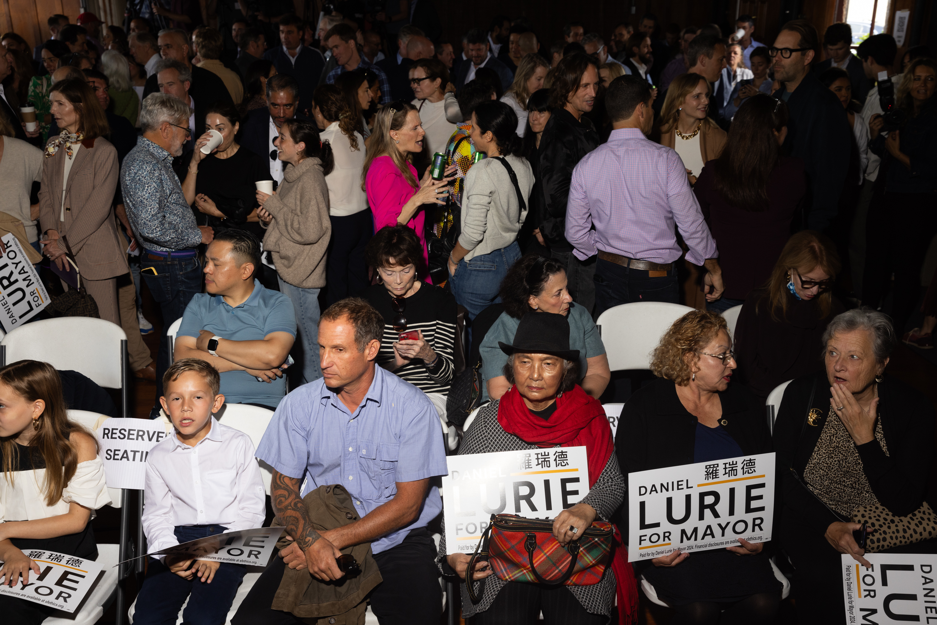 A diverse crowd holding &quot;Daniel Lurie for Mayor&quot; signs awaits an event in a packed hall.