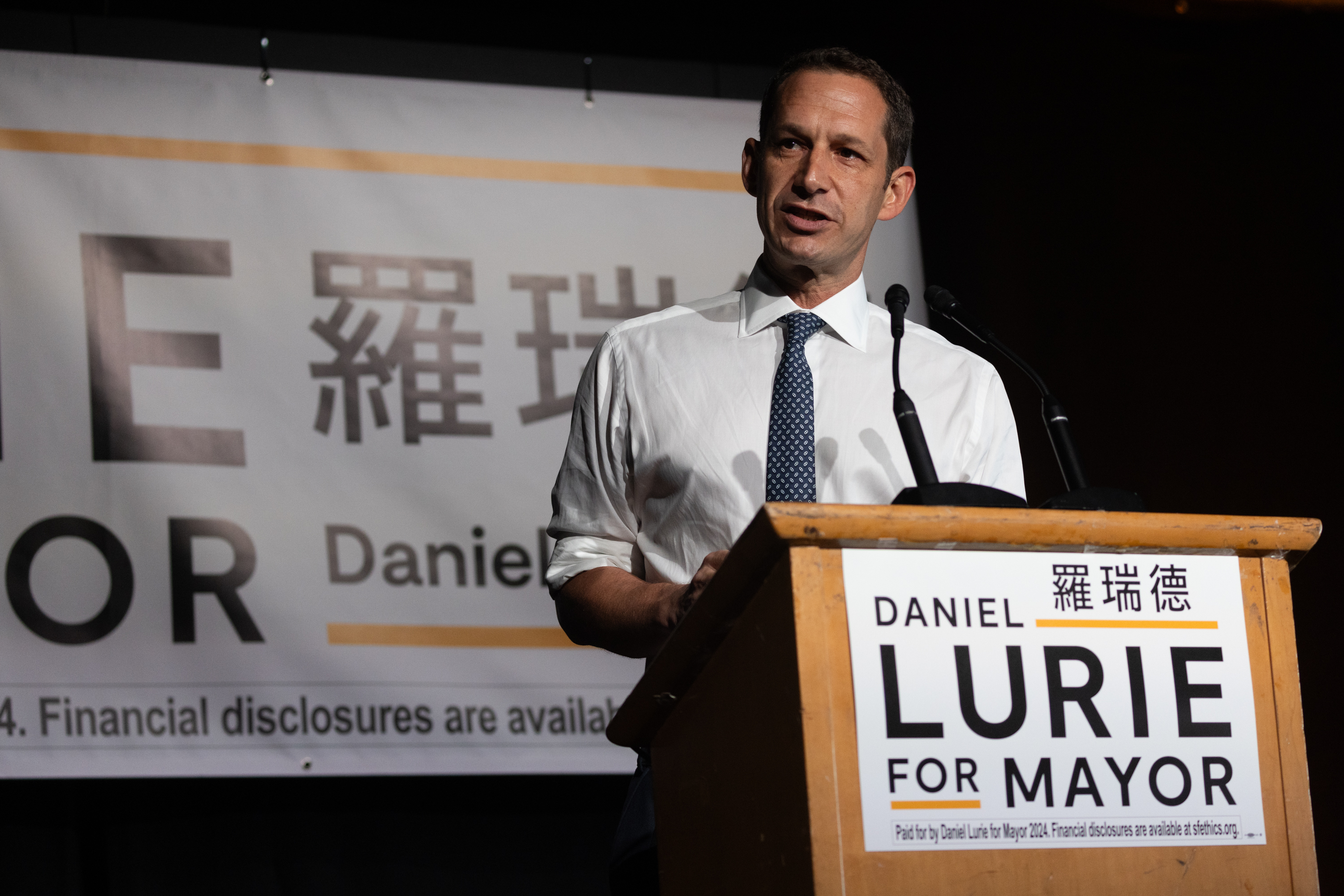 A man in a white shirt and tie speaks at a podium with "Daniel Lurie for Mayor" banners.