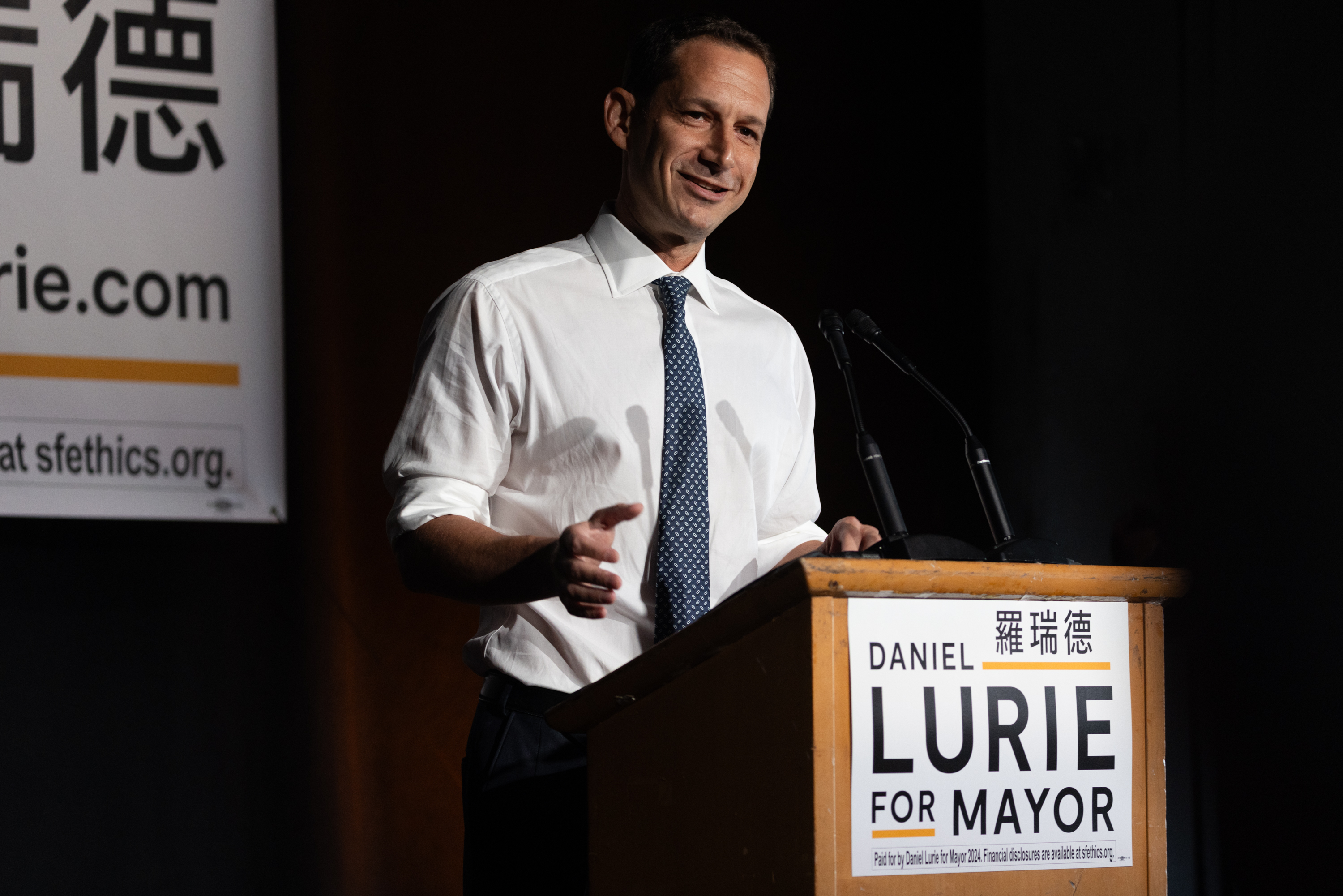 Daniel Lurie speaks at a podium with a &quot;Daniel Lurie for Mayor&quot; sign.