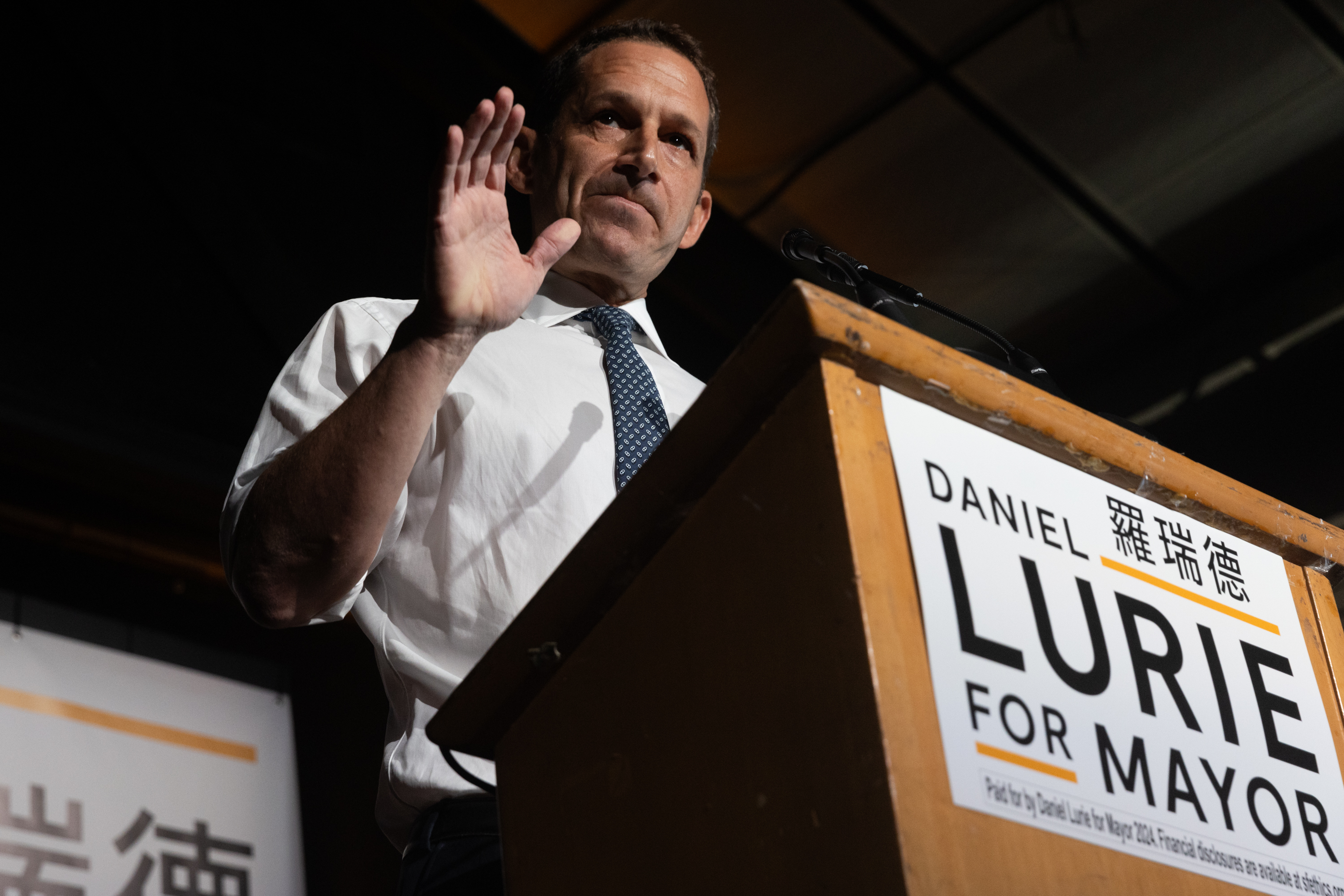 A man gestures while speaking at a podium with a "Lurie for Mayor" sign.