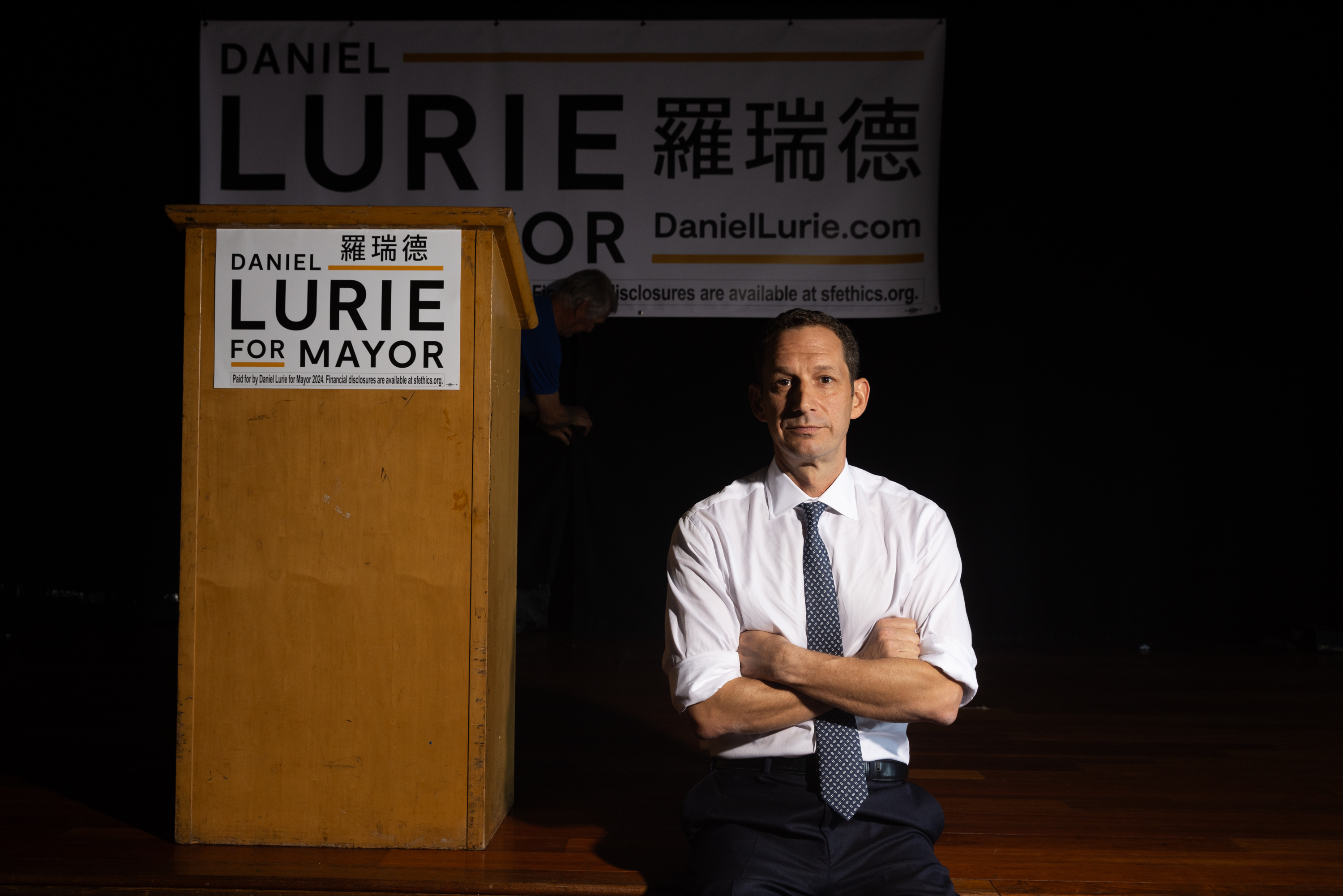 A man in a shirt and tie sits cross-armed near a podium with &quot;Daniel Lurie for Mayor&quot; signs.