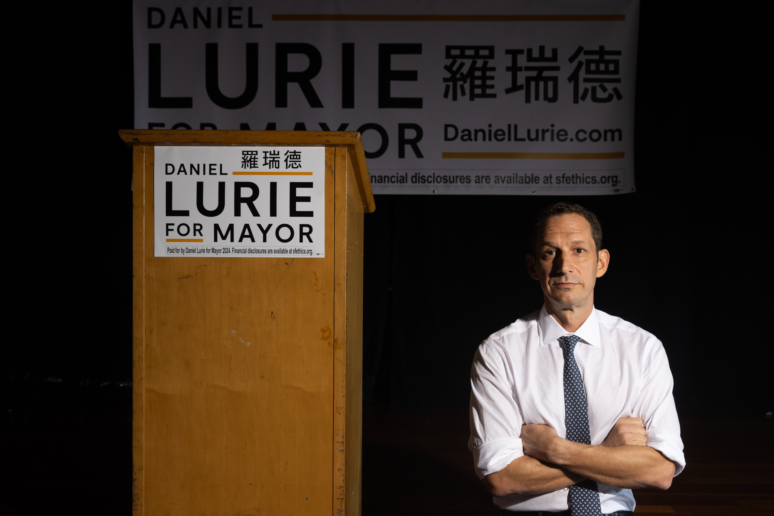 A man stands by a podium with "Daniel Lurie for Mayor" banners, arms crossed, looking forward.