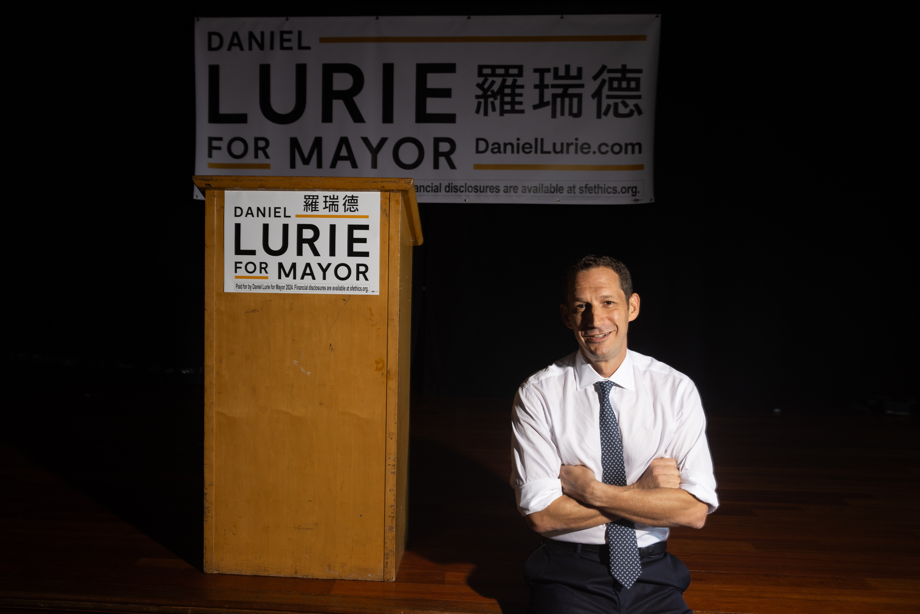 Man poses with arms crossed in front of sign Lurie for mayor