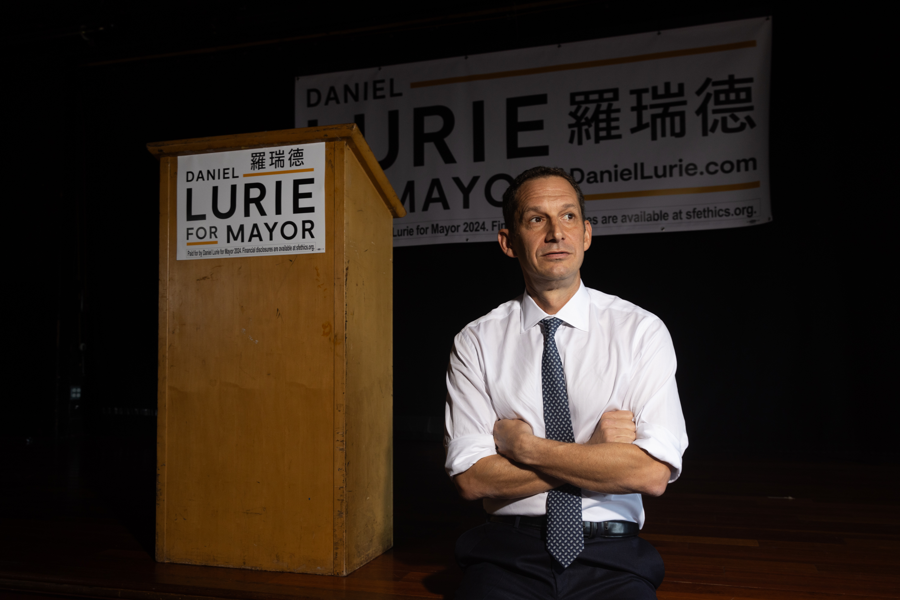 Daniel Lurie sits next to a podium with "Daniel Lurie for Mayor" signs, his arms crossed, wearing a shirt and tie.