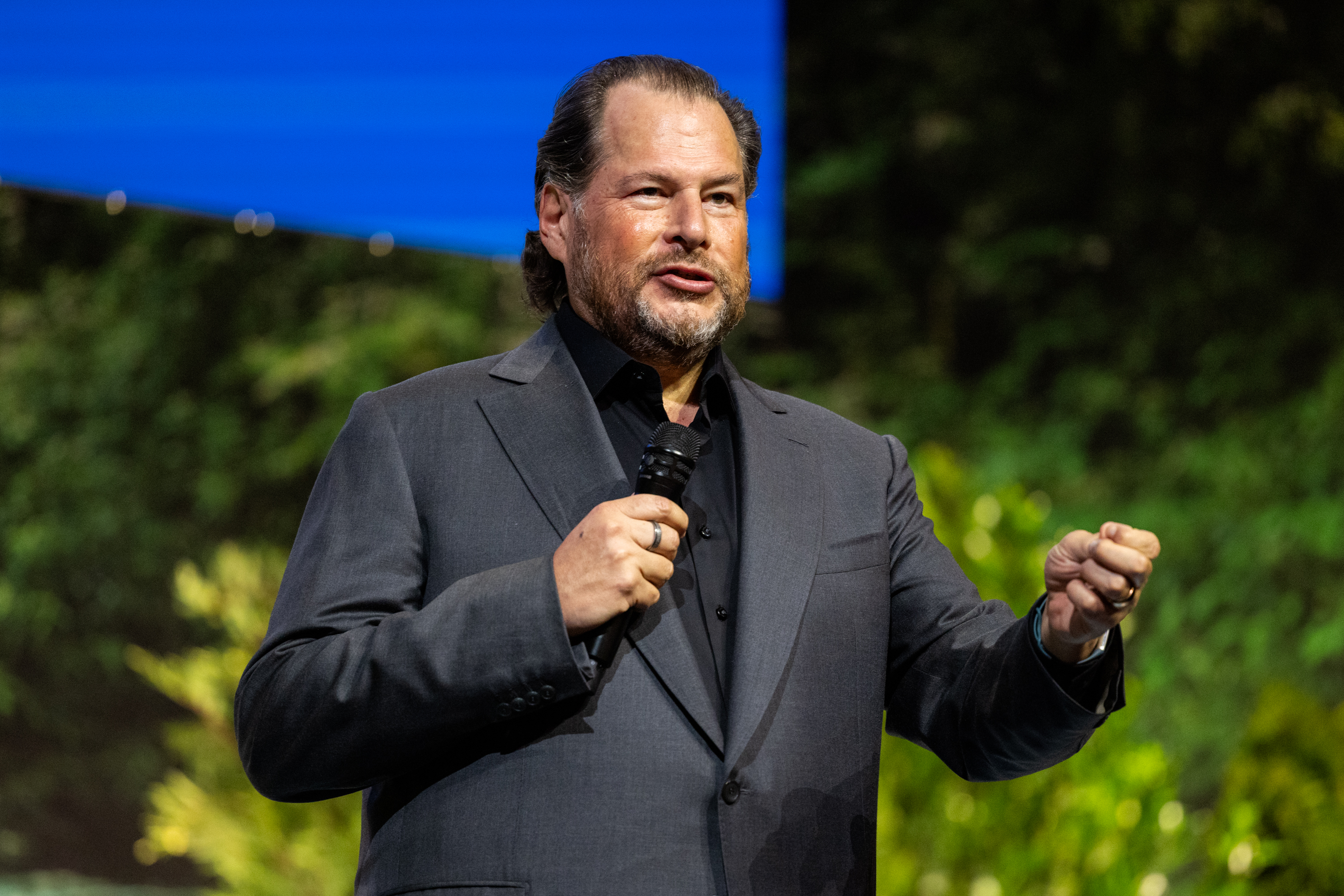 A man in a gray suit and black shirt is speaking into a microphone on stage, gesturing with one hand. The background is a mix of green foliage and a blue screen.