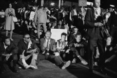 A black &amp; white photo of people seated on a road, displaying protest signs.