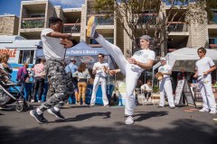 People perform Capoeira, a Brazilian martial art, on a sunny street with onlookers and a library mobile in the background.