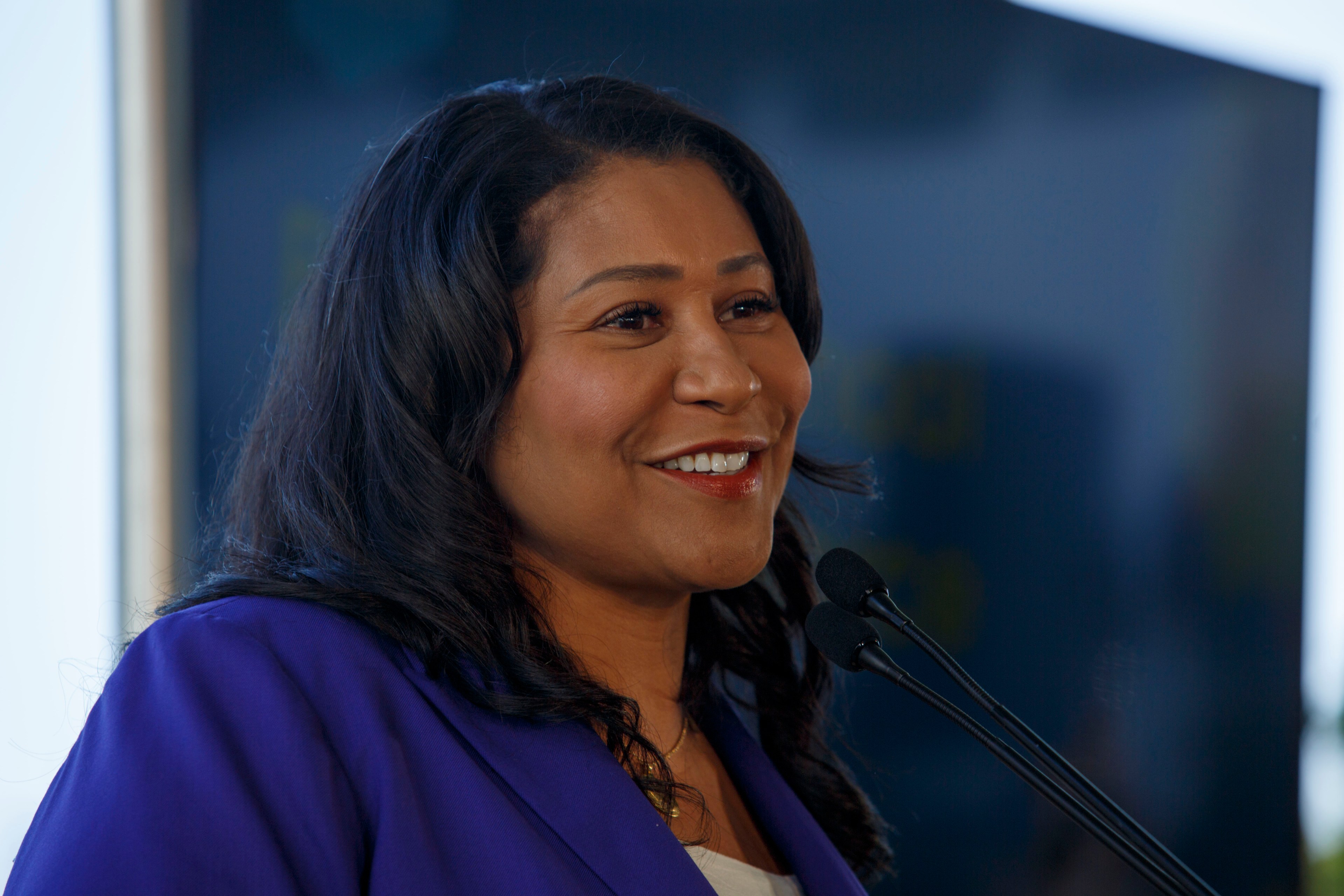 A woman with long, dark hair is smiling and speaking at a podium with microphones, wearing a purple blazer against a blurred dark background.