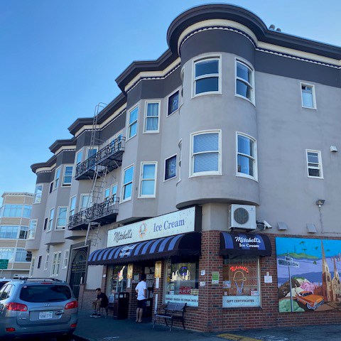 The image shows a beige, multi-story building with an ice cream shop on the ground floor. The shop has a sign and a striped awning, with a mural painted on the side wall.