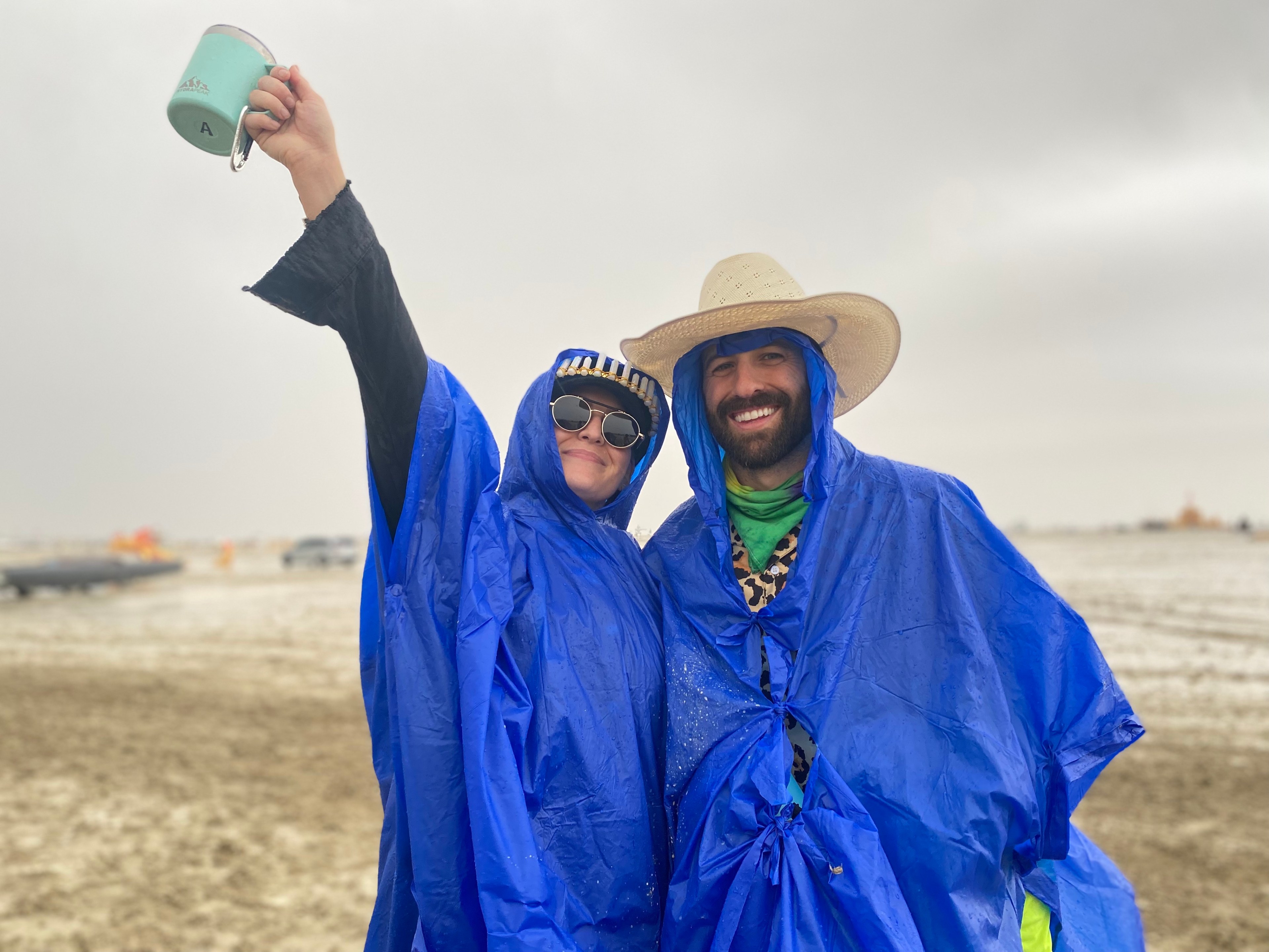 Two people in blue ponchos are smiling; one is raising a mug, the other wearing a straw hat.