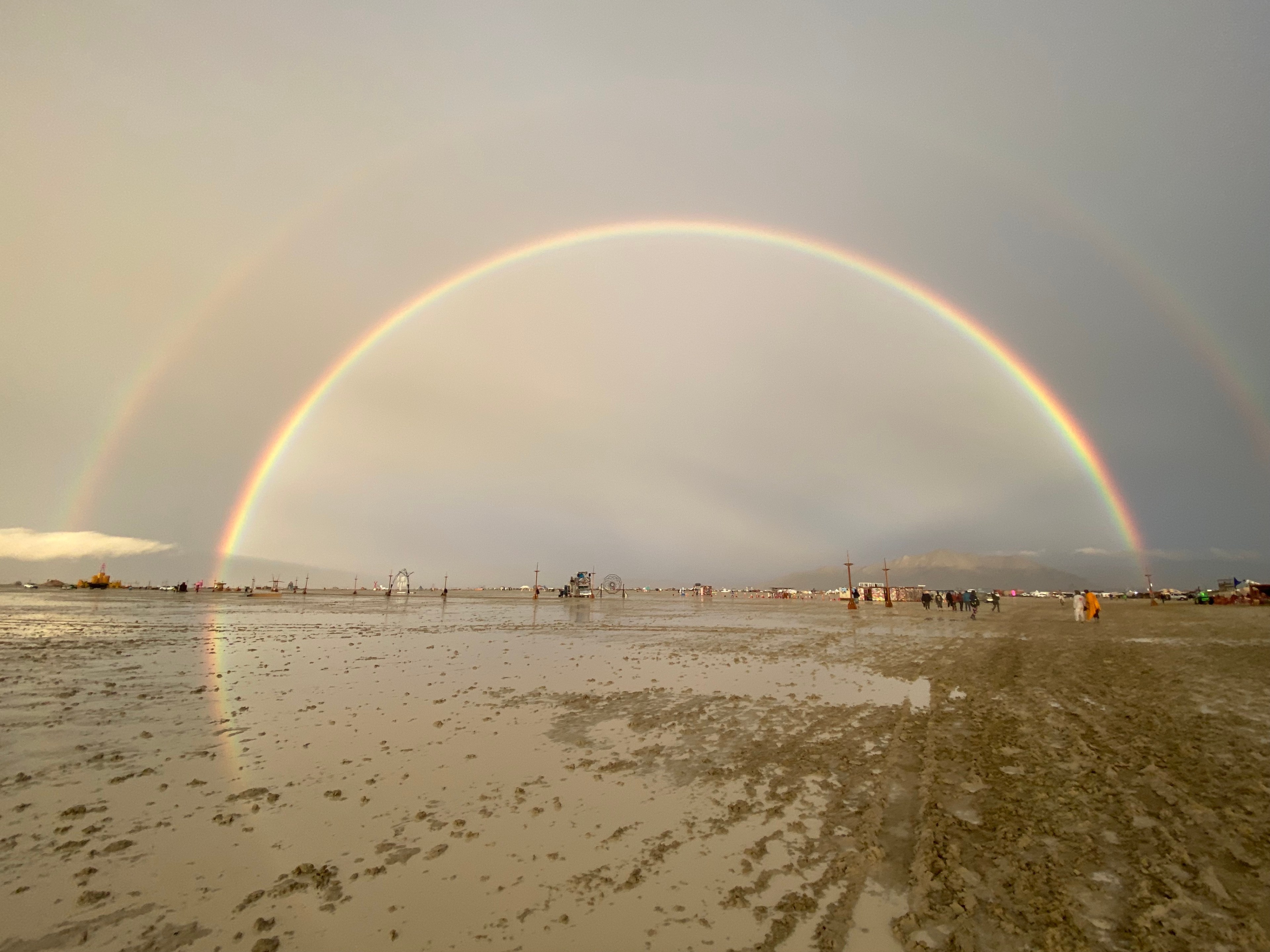 A double rainbow arches over a wet, muddy landscape with scattered people and structures.
