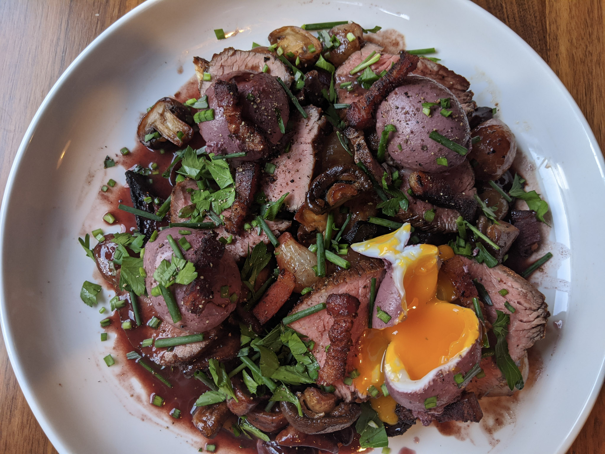 A plate of seared tri-tip steak and eggs en meurette poached in red wine with roasted mushrooms, onions and bacon rests on a wide white plate.